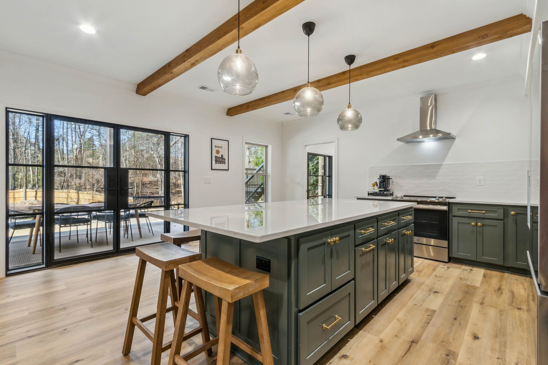 Modern kitchen with green cabinets, white countertop island, wooden beams, and large windows overlooking a patio.