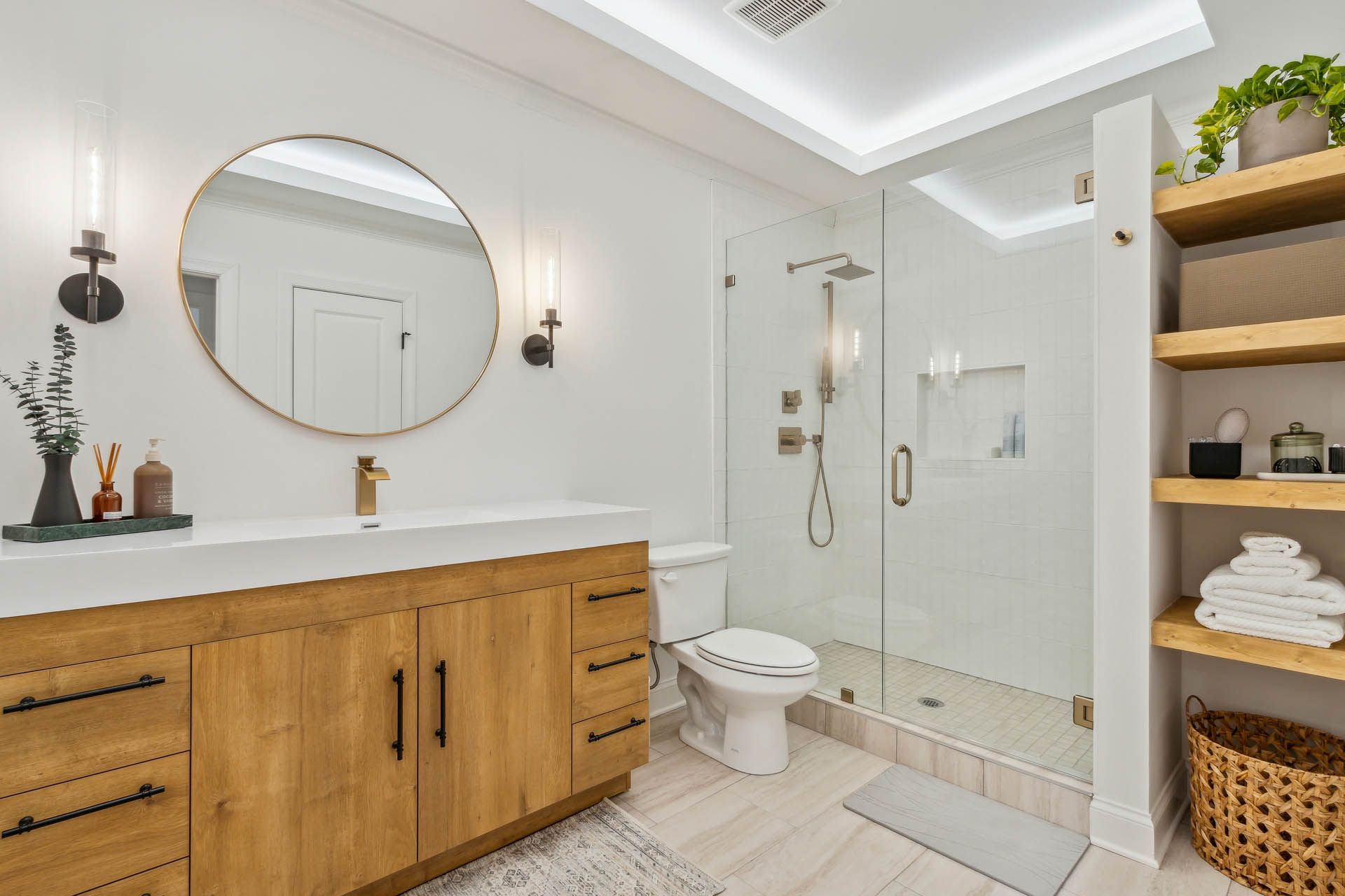 Bathroom with a light wood vanity, round mirror, glass shower, and wooden shelves. White walls and flooring.