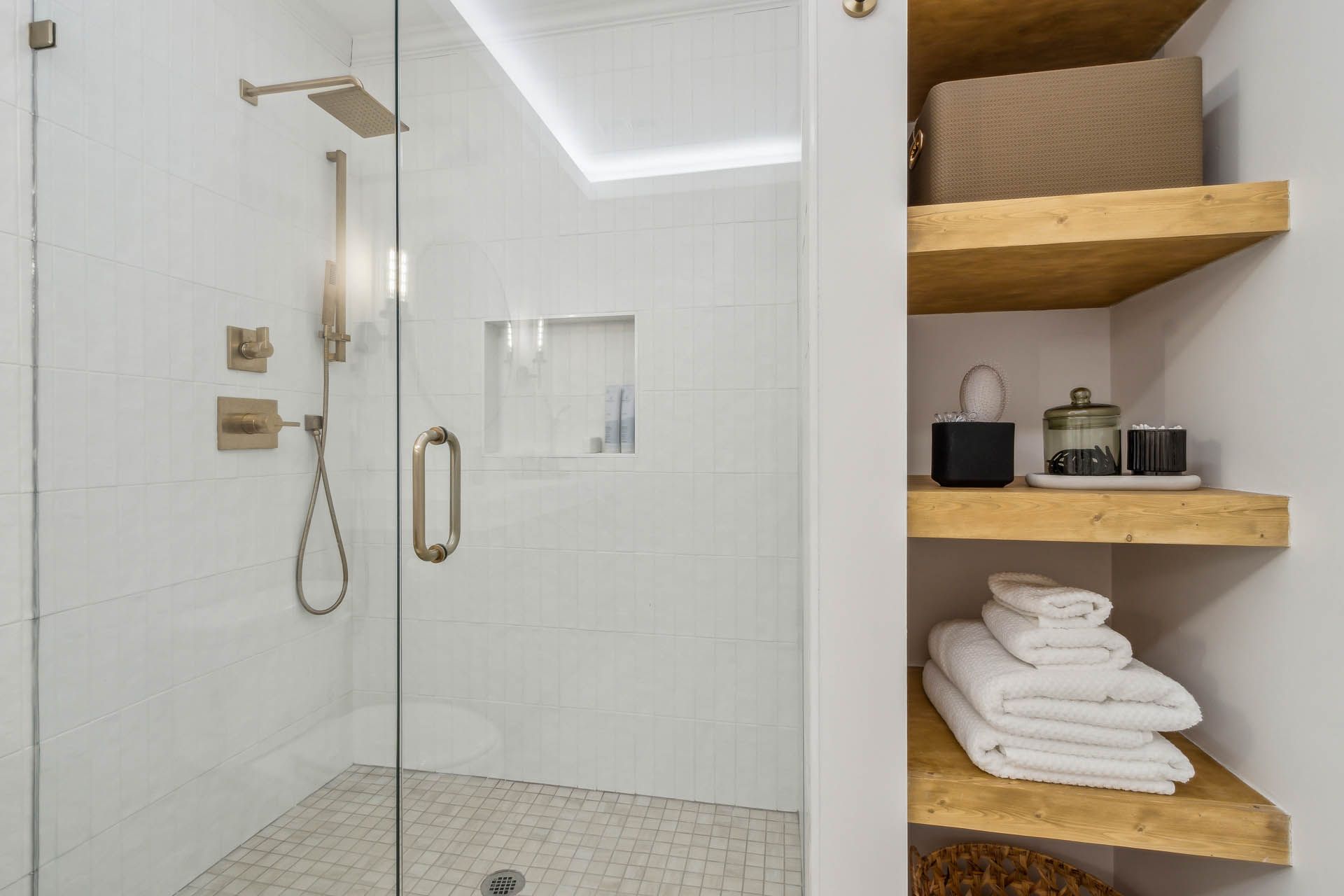 Modern white bathroom with a glass shower, brass fixtures, and wood shelving.