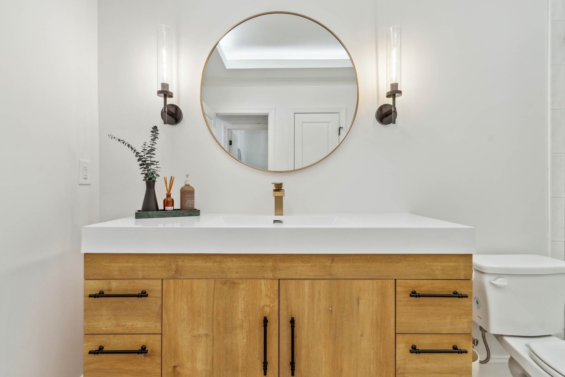 Bathroom with light wood vanity, round mirror, gold faucet, and sconces.