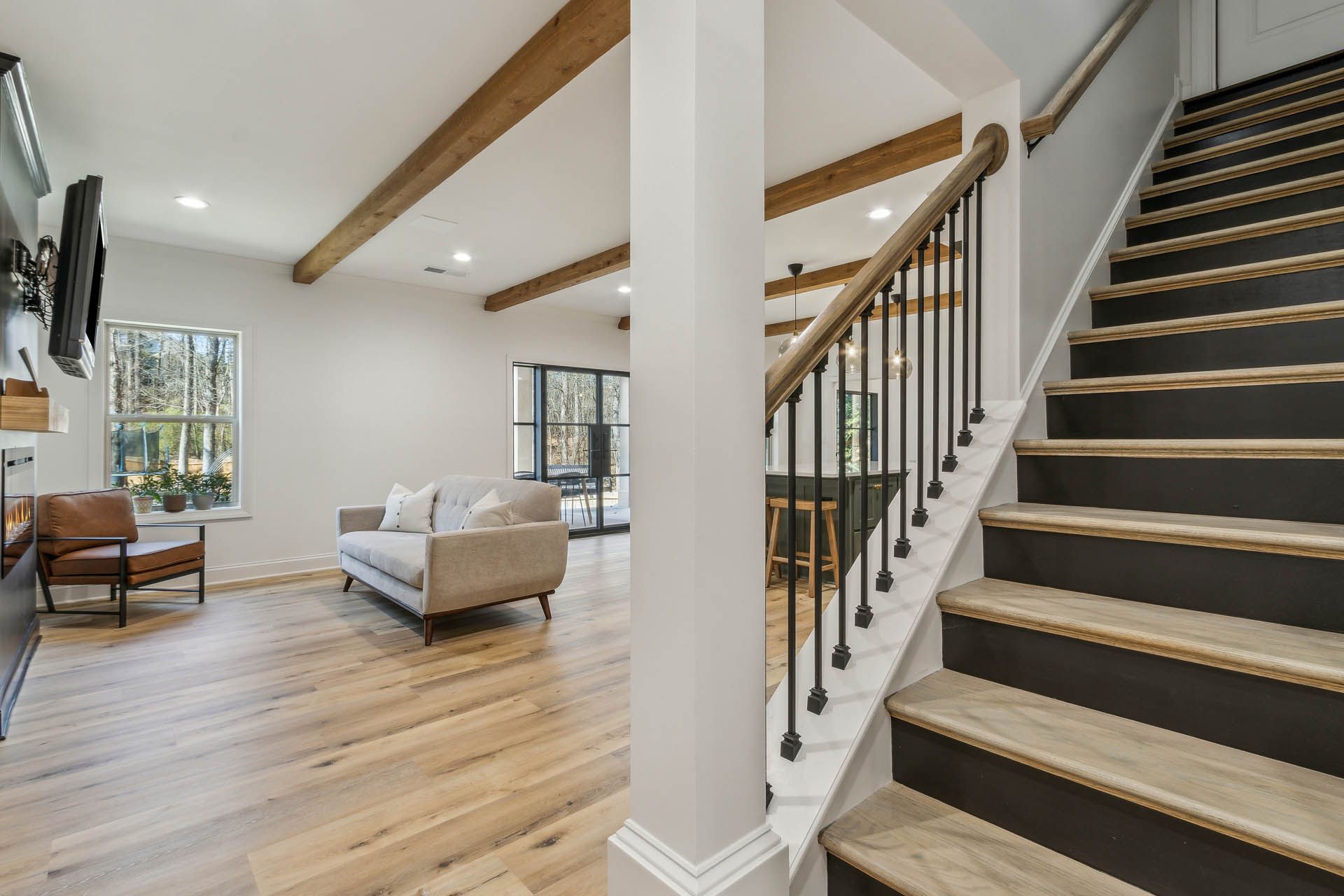 Interior view of a modern home with a staircase, living area, and wooden beams. The walls are white, and the floors are light wood.