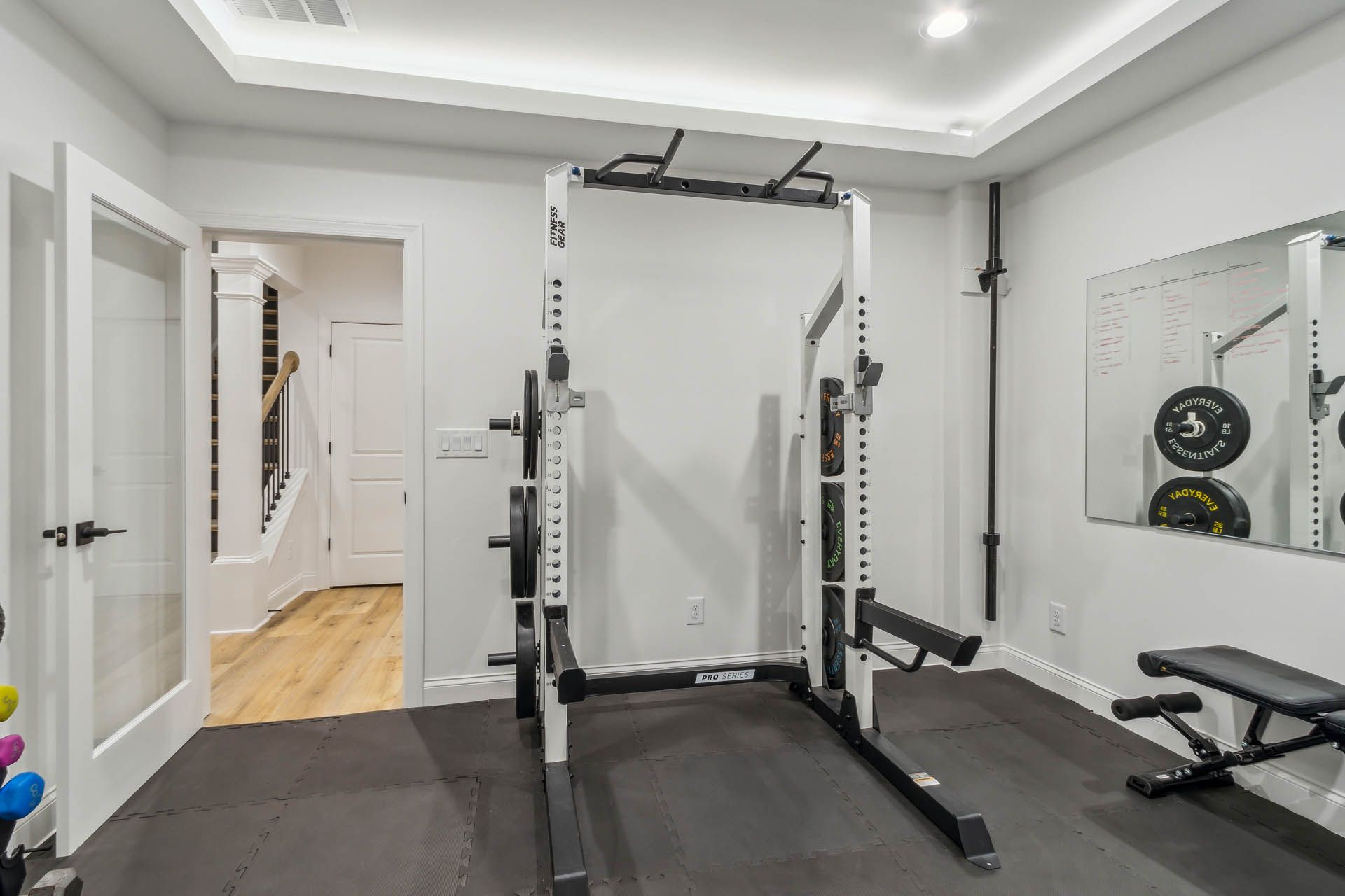 A home gym with a squat rack, weight bench, and mirror.  The room has gray walls and black flooring. A doorway leads to stairs.