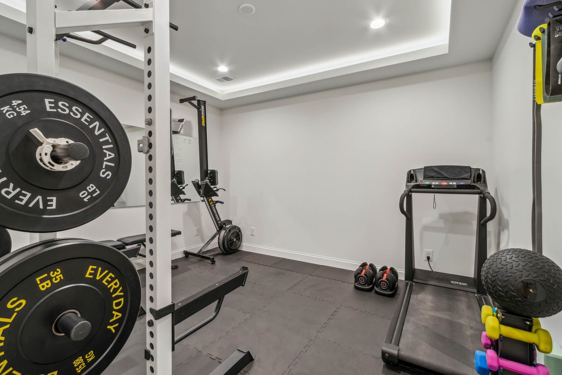 A home gym with a squat rack, rowing machine, treadmill, weights, and a weight bench on a black rubber floor.
