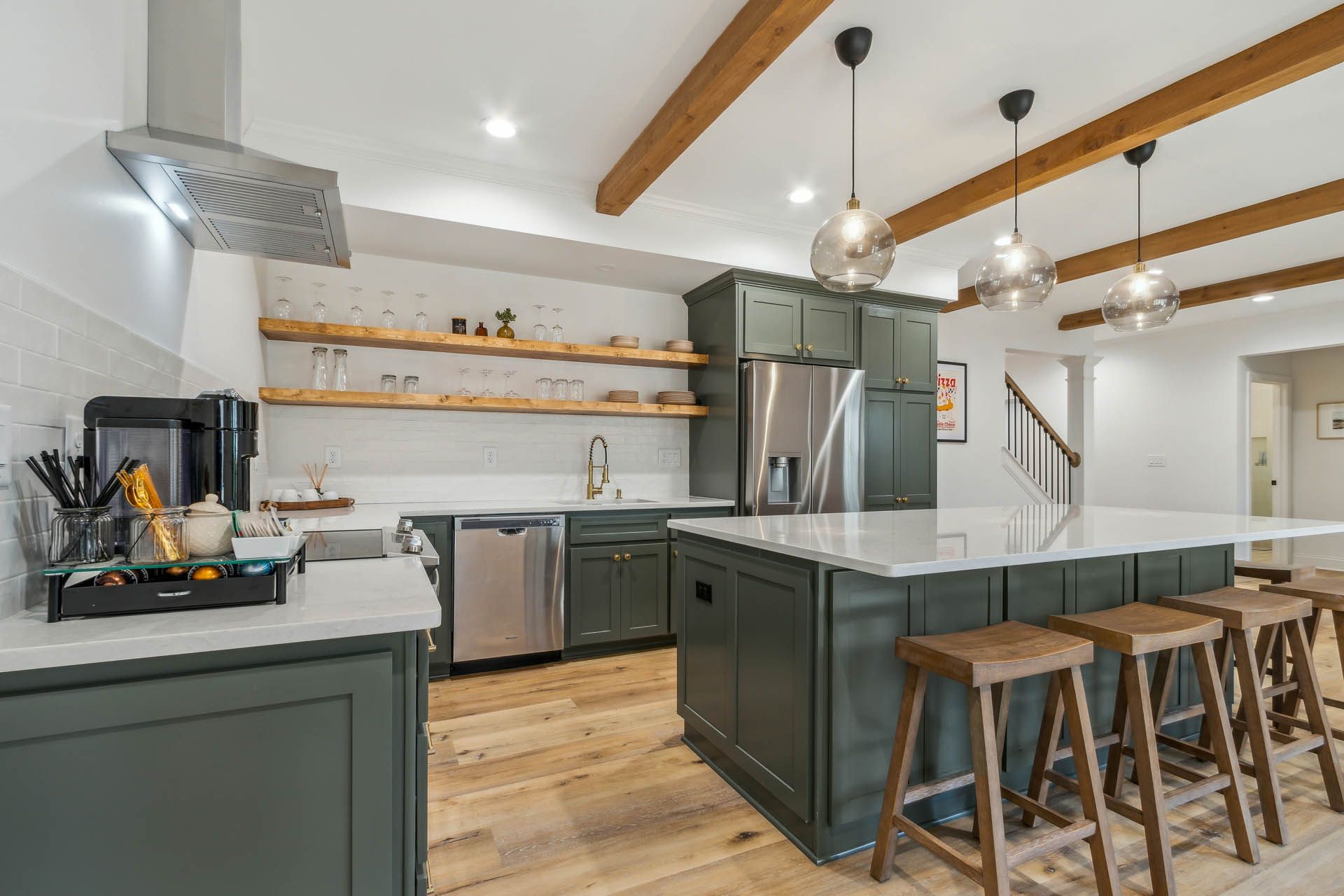 A modern kitchen with green cabinets, stainless steel appliances, and a large island with stools. Wooden beams and shelves add warmth.