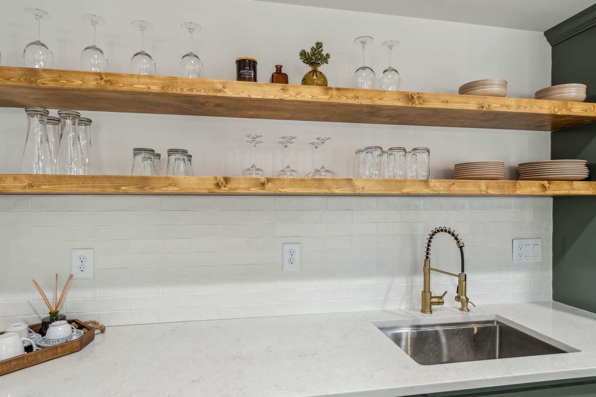 A kitchen sink with wooden shelves holding glassware. The countertop is white, and the wall has white subway tiles.