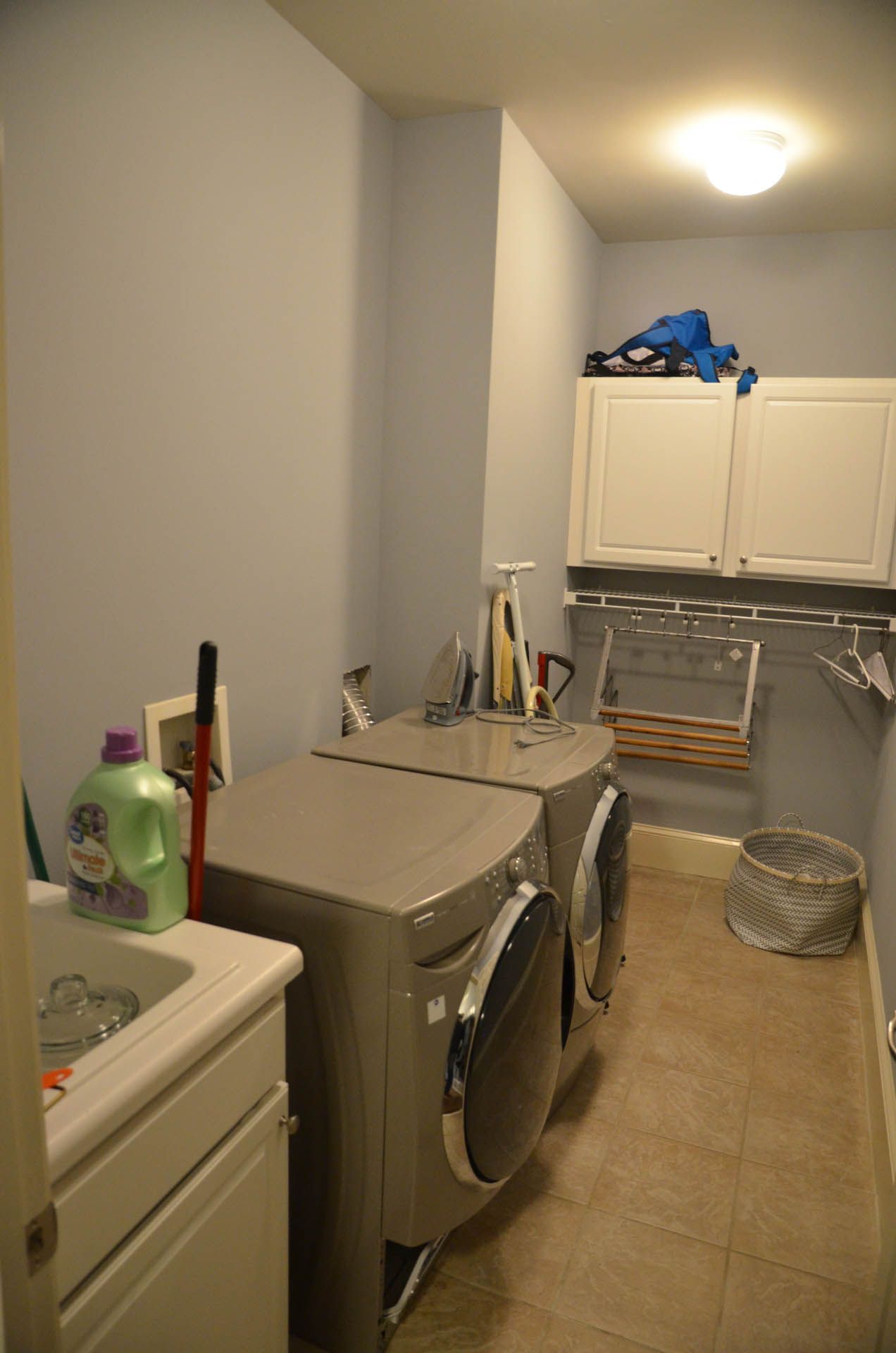 A laundry room with a washing machine, dryer, cabinets, a sink, and drying rack. The walls are light blue.