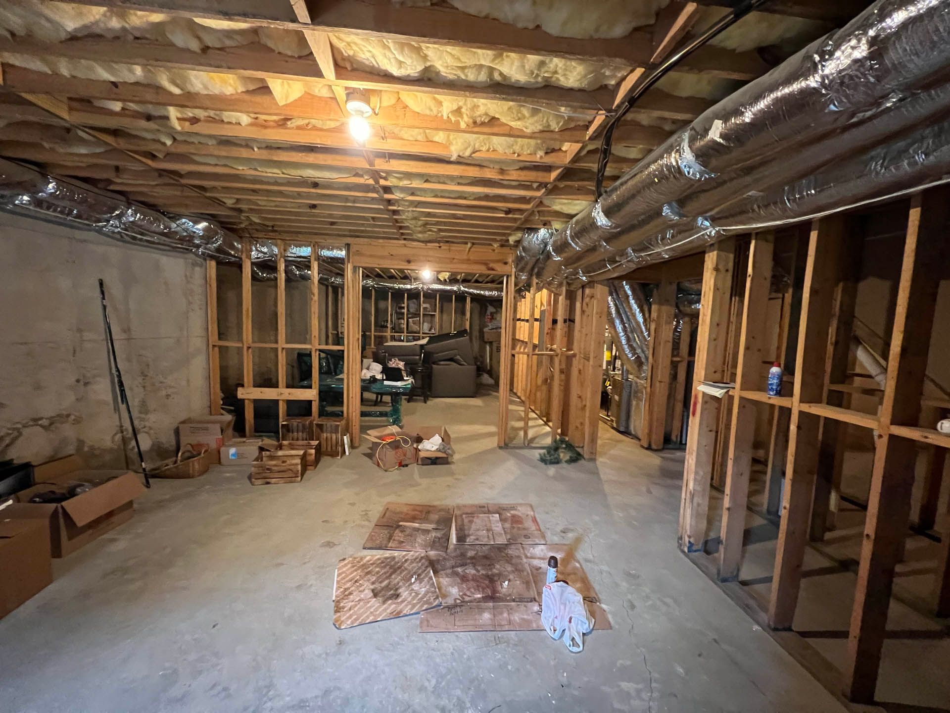 Basement under construction with exposed wooden framing and ductwork overhead. Concrete floor, insulation, and scattered debris are visible.