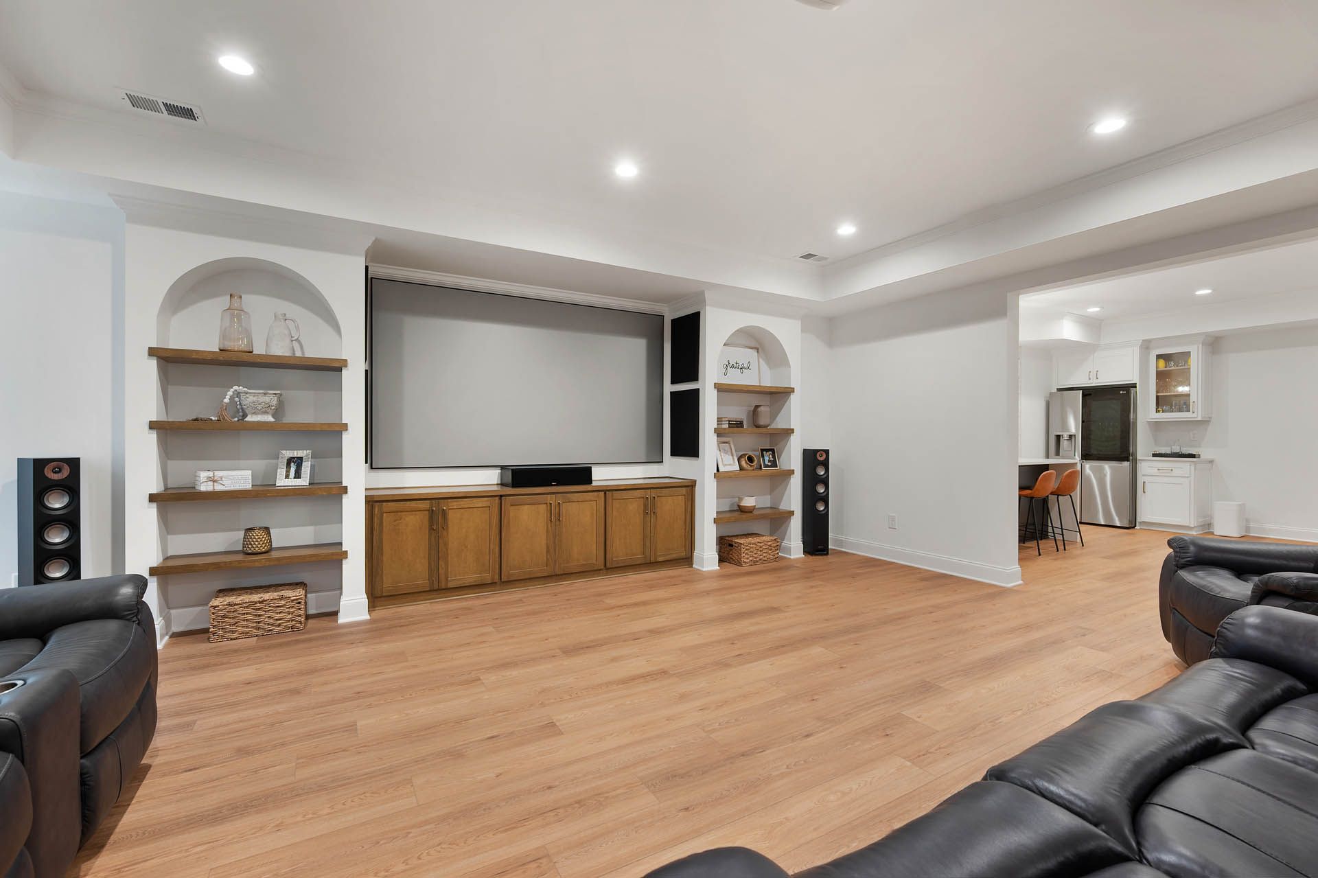 A home theater room with hardwood floors, built-in shelving, and a large screen. The room has a wooden cabinet under the screen and a black leather couch.