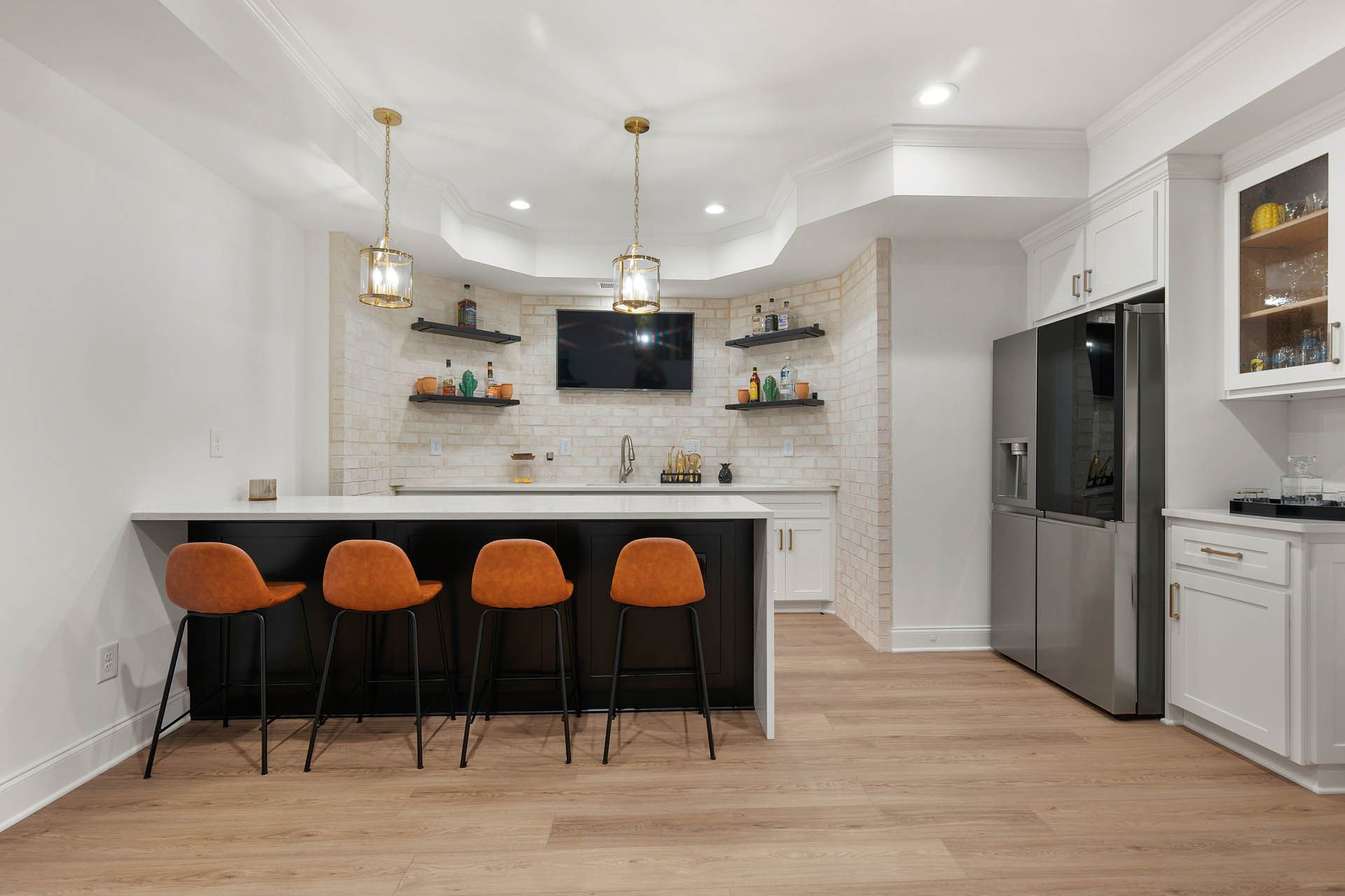 A modern basement bar area featuring a black and white counter with orange bar stools, a stainless steel refrigerator, and a small TV.