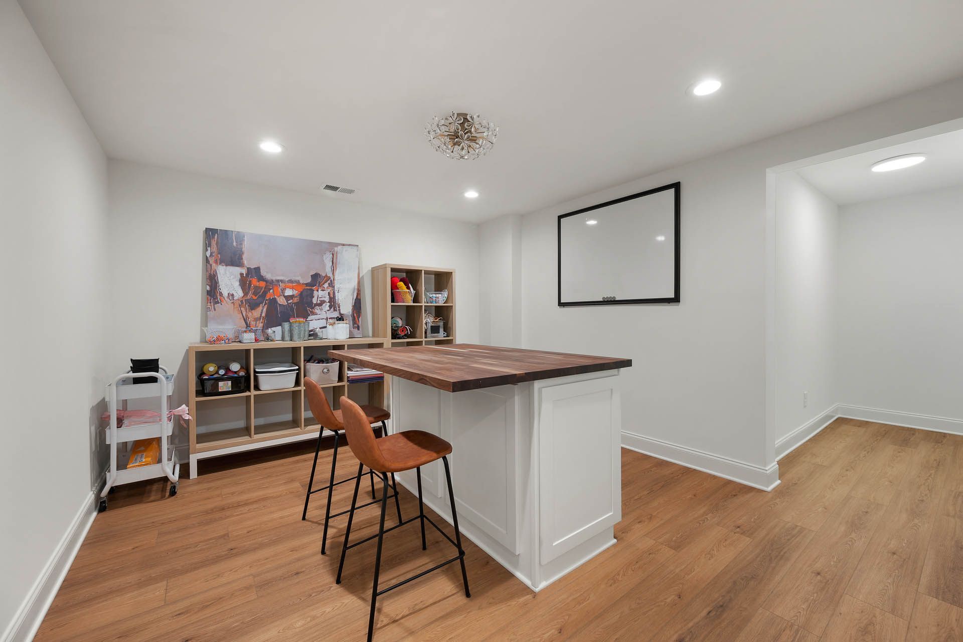A finished basement with a bar and two brown stools, wood floors, built-in shelves with snacks, and a large family portrait.