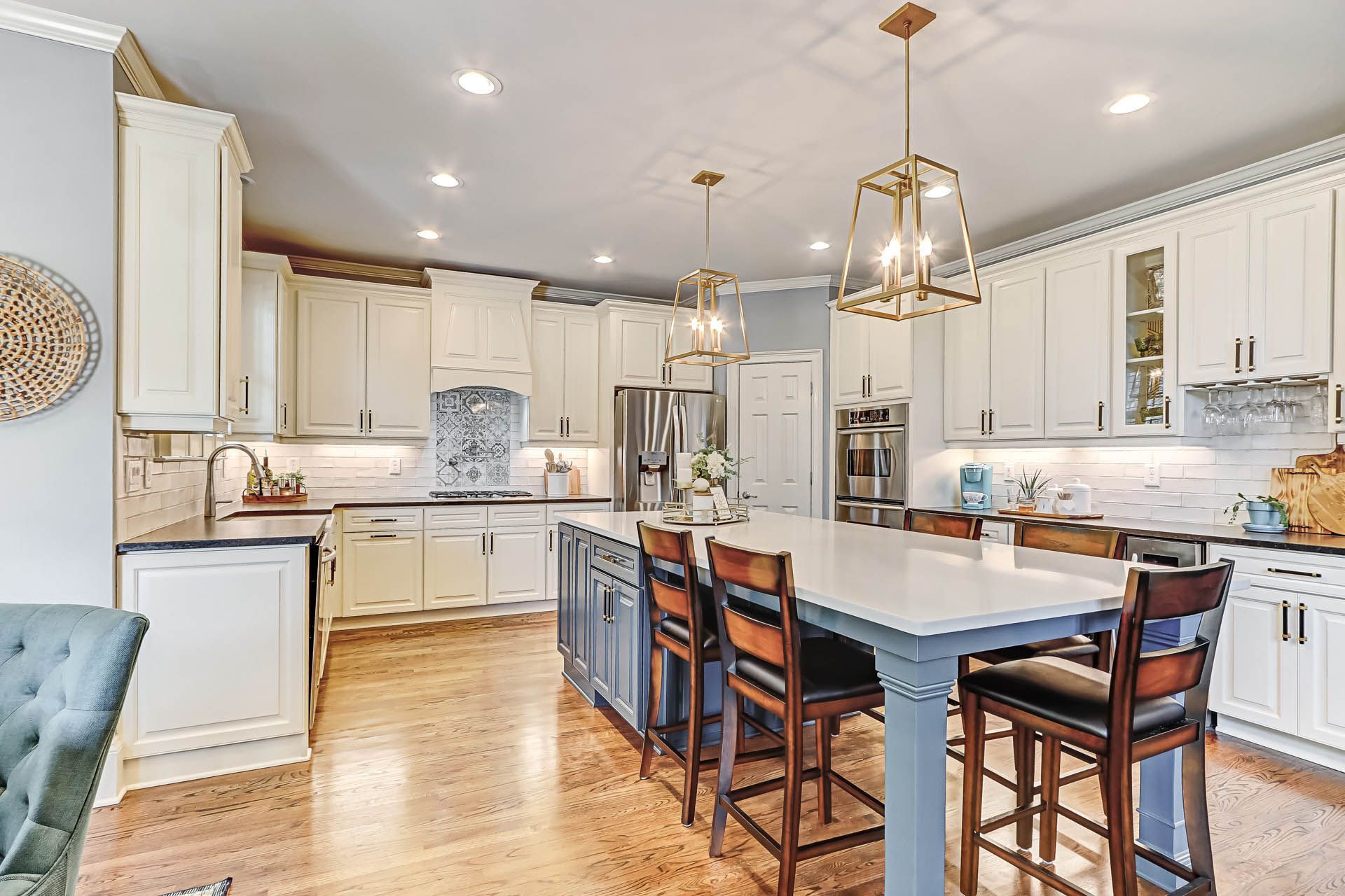 Bright kitchen with cream cabinets, blue island, wood floors, and gold light fixtures. Bar stools are placed around the island.