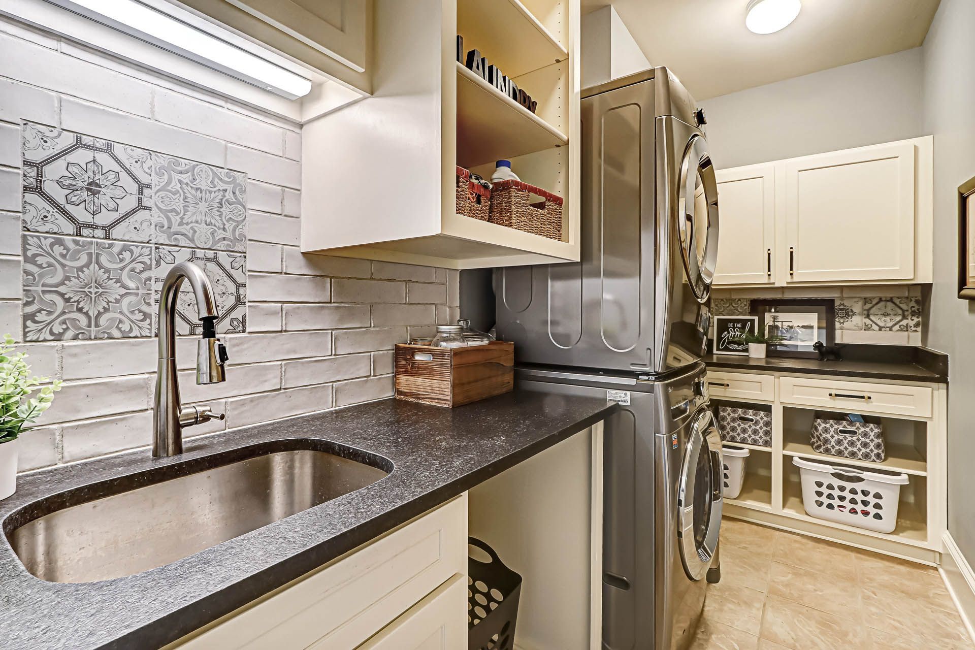 Laundry room with a sink, countertop, and stacked washer and dryer. Light-colored cabinets, patterned backsplash, and baskets.