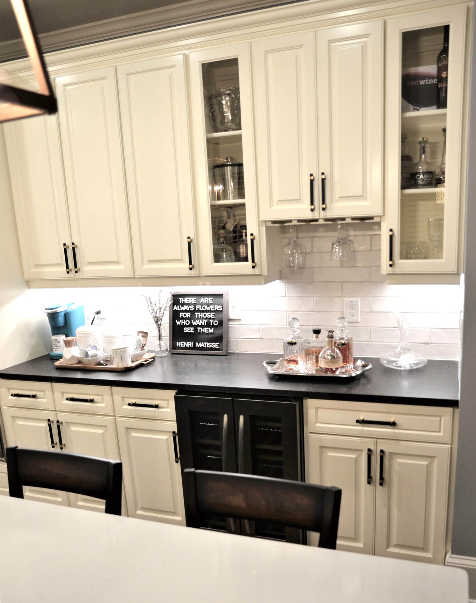 Cream-colored kitchen bar with a wine fridge, cabinets, and countertop, adorned with glasses and bottles.