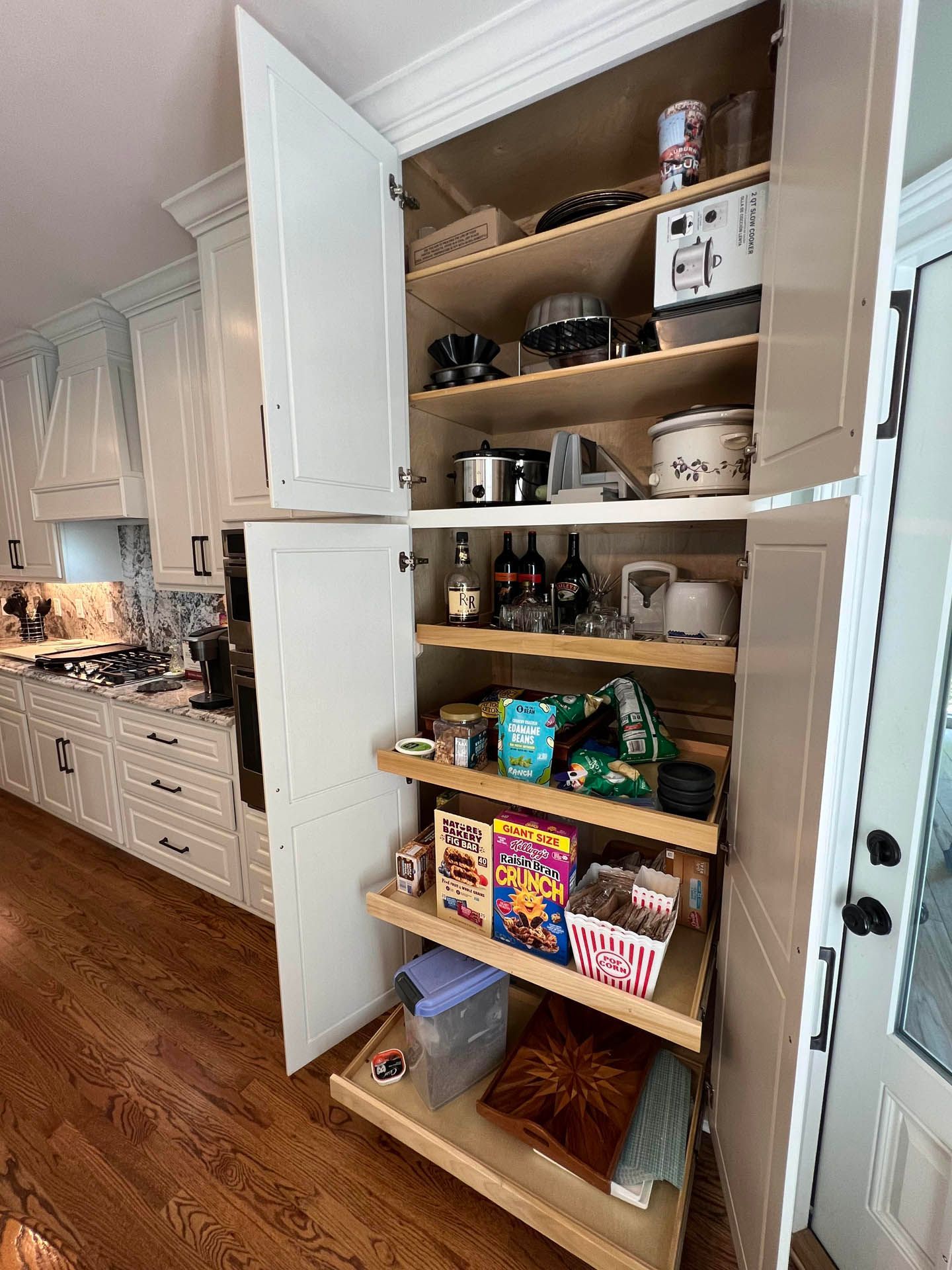 White kitchen pantry cabinet with open doors revealing pull-out shelves filled with food and appliances.