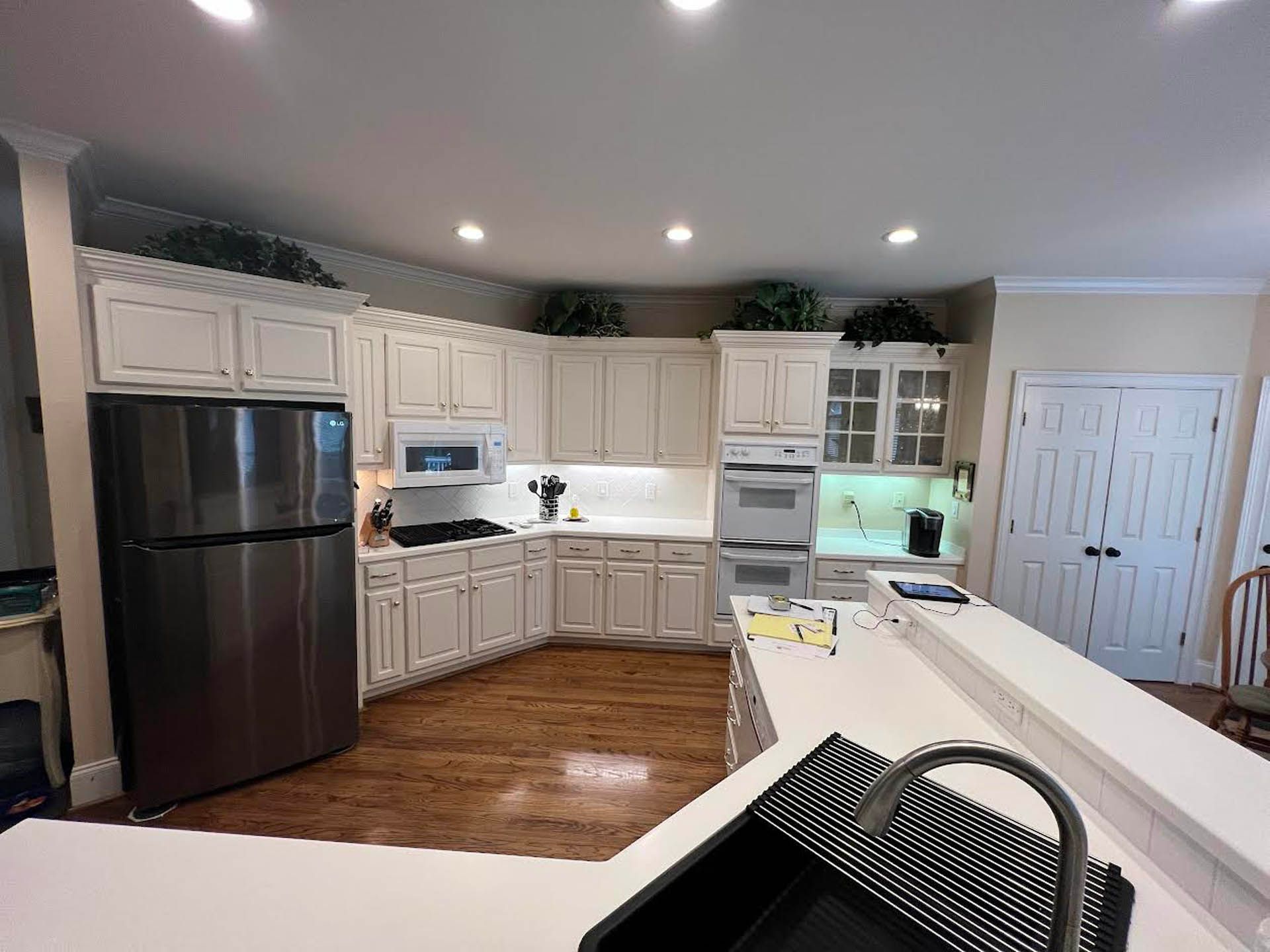 A kitchen with white cabinets, stainless steel refrigerator, and a dark sink.  Light wooden floors and a white countertop are also present.