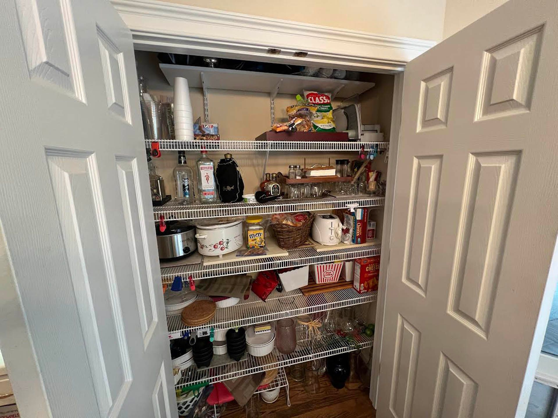 A pantry with white wire shelves filled with various kitchen items, including appliances, food, and glasses. The doors are white and open.