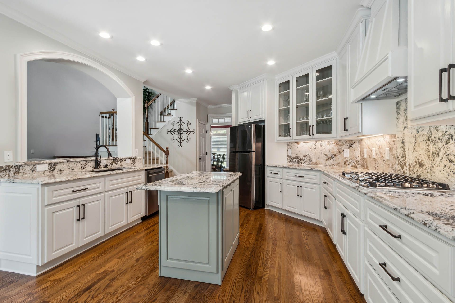 A bright, white kitchen with a central island and granite countertops, leading to a staircase and entryway.