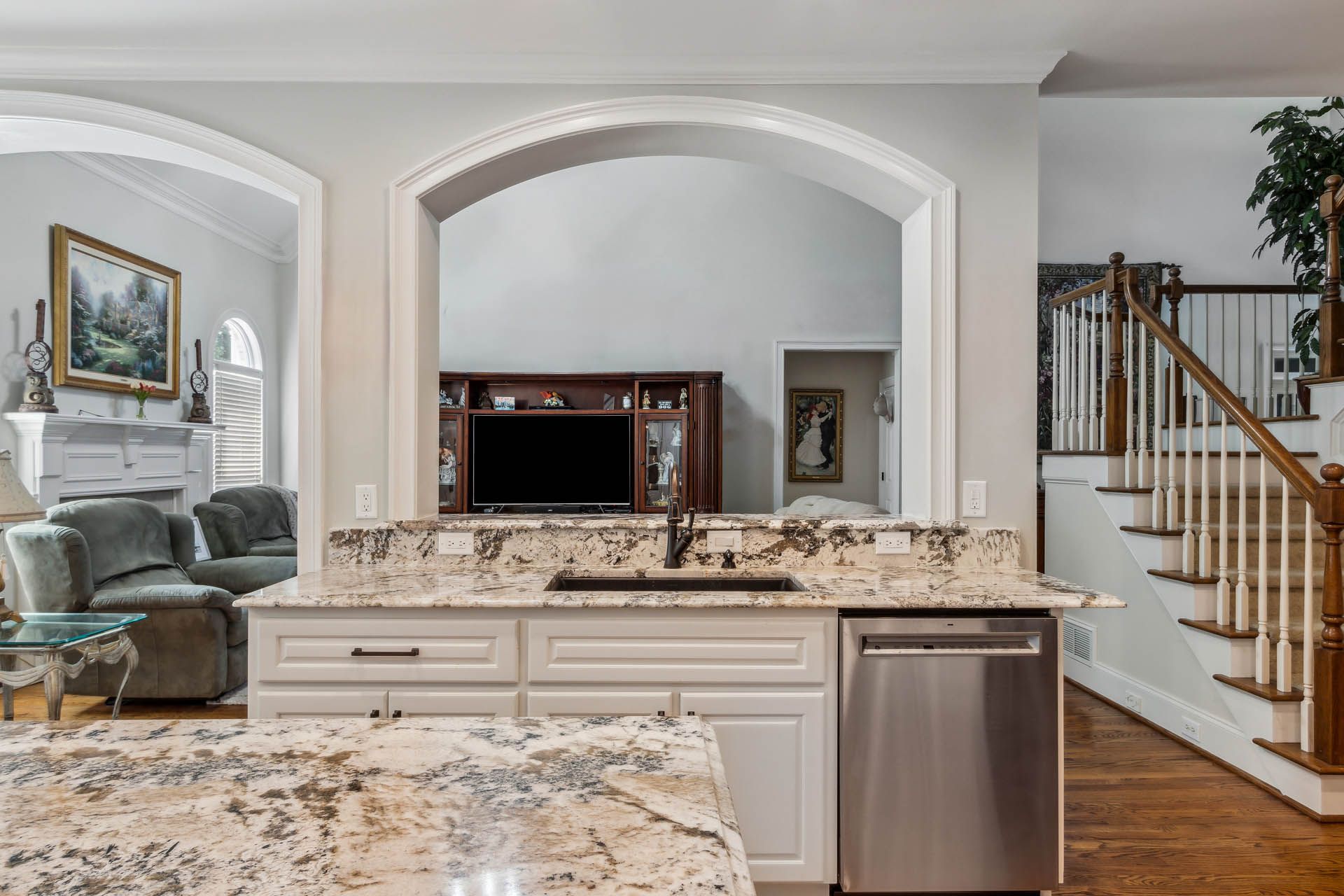 Kitchen with a granite countertop island, stainless steel dishwasher, and view of a living room and staircase.