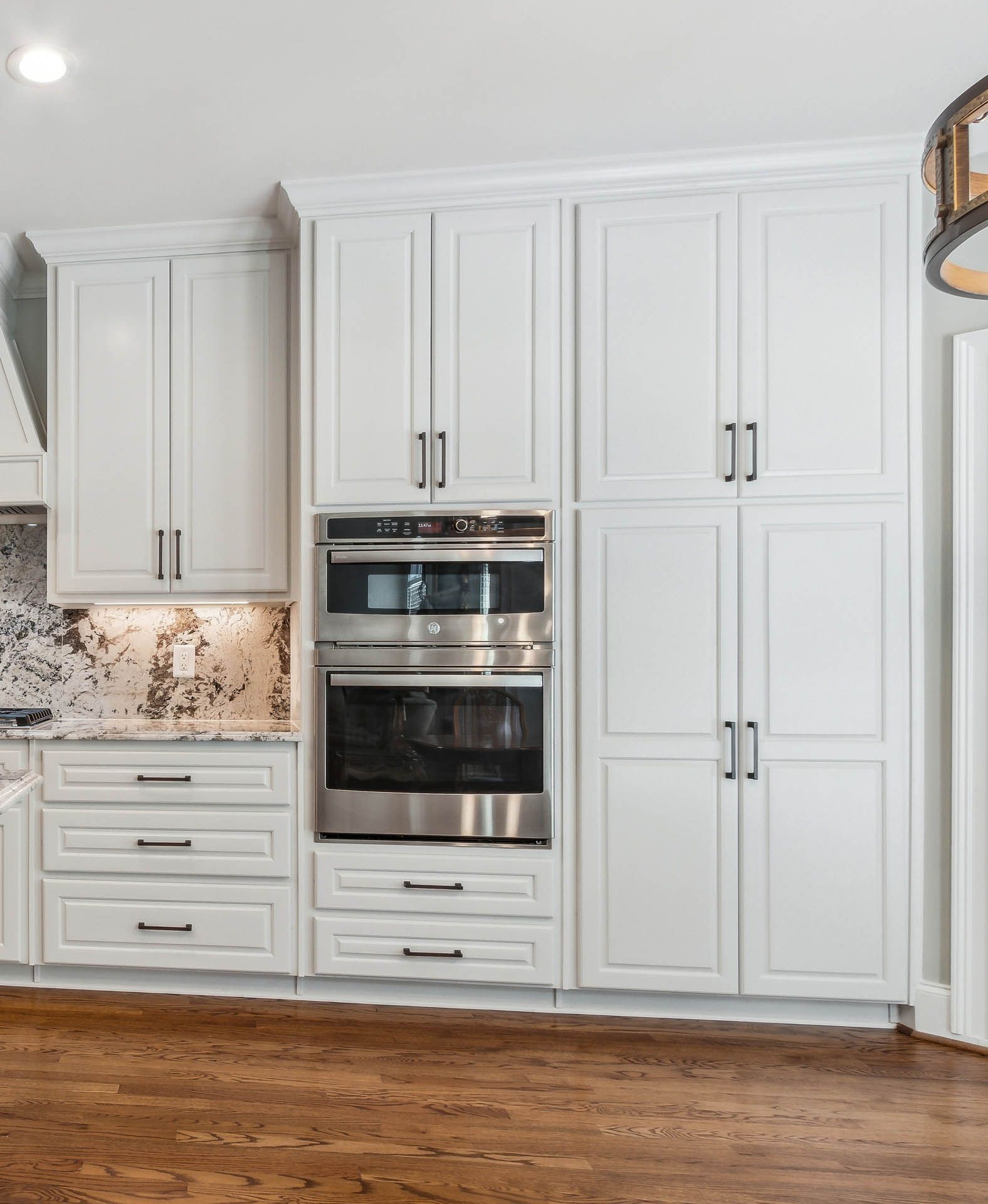 White kitchen cabinets with a built-in double oven and black hardware, set against a granite countertop and hardwood floor.