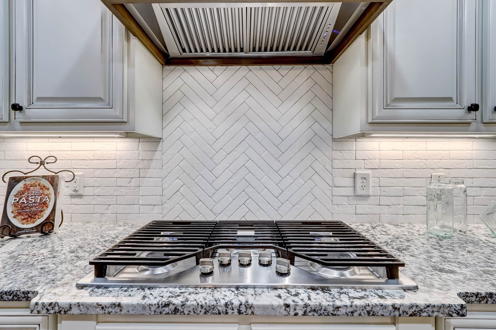 Kitchen stove with a white herringbone tile backsplash, beneath a range hood, granite countertop, and white cabinets.