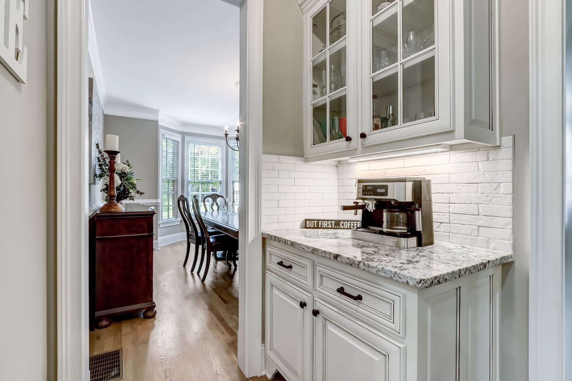 A white-cabineted coffee station with a granite countertop, a coffee maker, and a white-tiled backsplash. It is situated in a hallway.