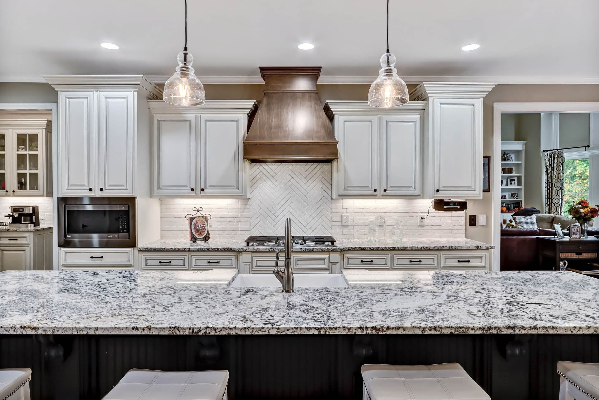 A modern kitchen featuring white cabinets, a dark wood range hood, and a granite countertop island. Two pendant lights hang overhead.