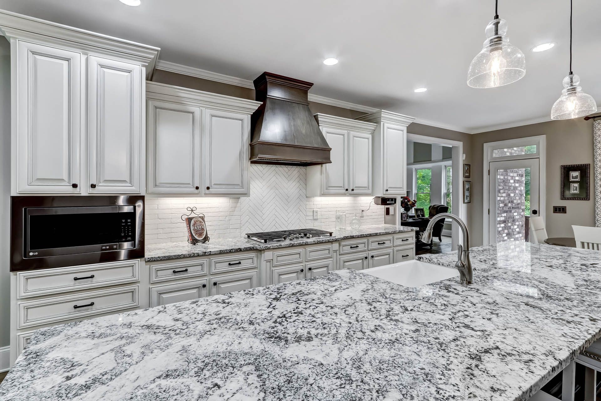 A modern kitchen with white cabinets, granite countertops, stainless steel range hood, and a view to the outside.