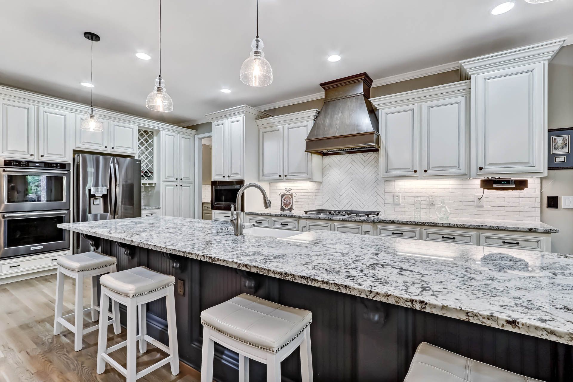A modern kitchen with white cabinets, a large granite island with seating, stainless steel appliances, and pendant lighting.