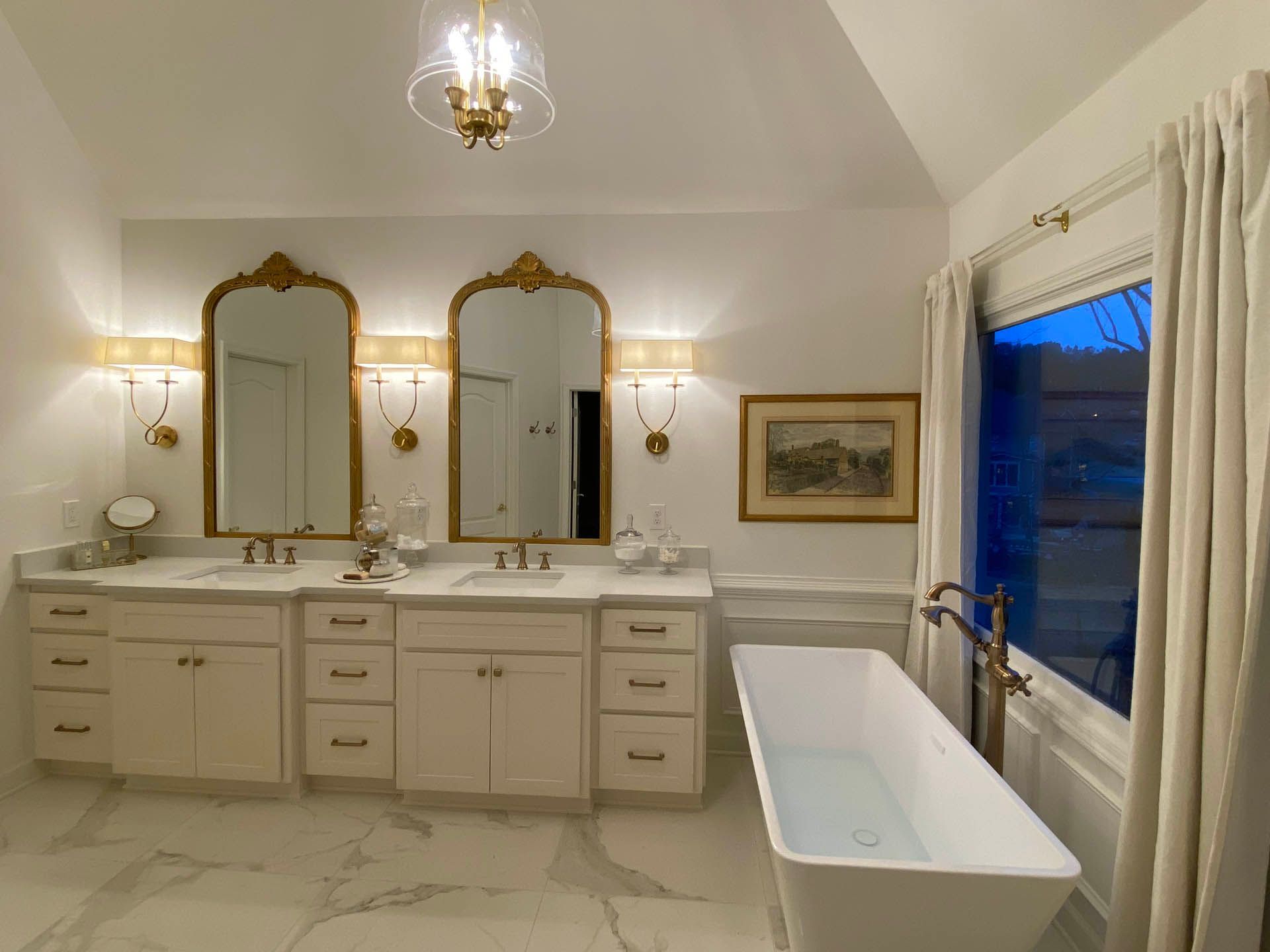 Elegant white bathroom with double vanity, gold mirrors, and a freestanding tub. Soft lighting and a framed picture complete the luxurious space.