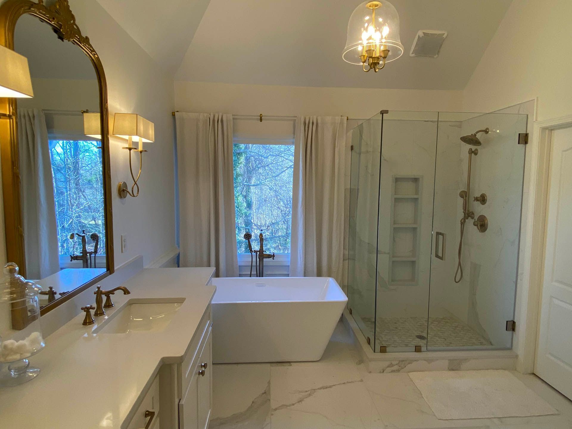 Bathroom with white walls, marble floor, glass shower, and a freestanding tub near a window with curtains. Gold-framed mirror and fixtures.