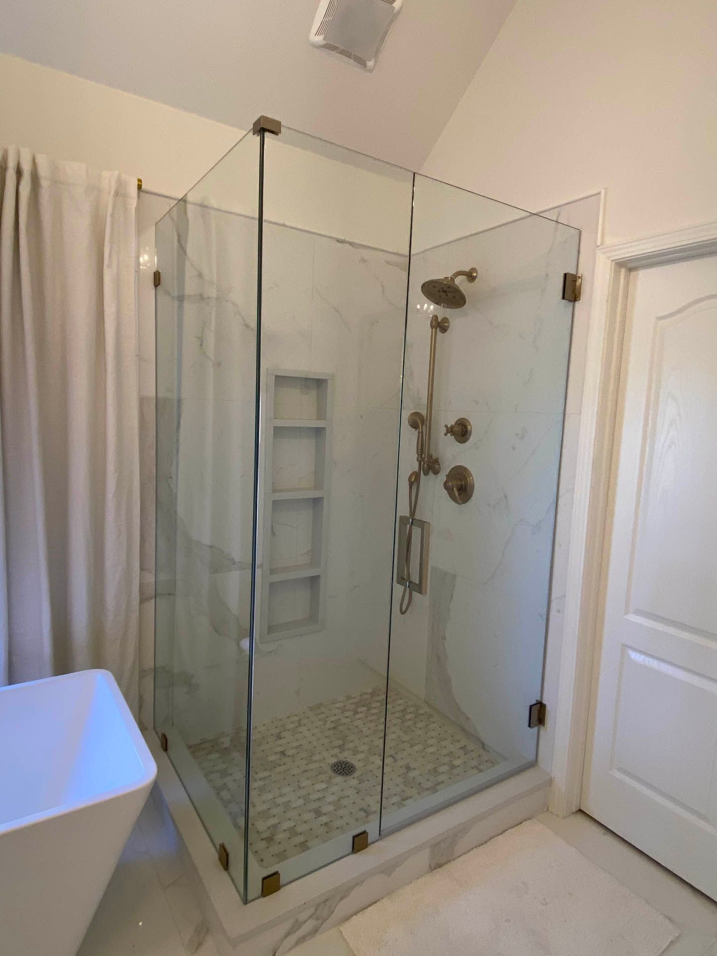 A luxurious glass shower enclosure with gold-colored hardware. The interior features a stone floor, built-in shelving, and a marble-like tiled wall.