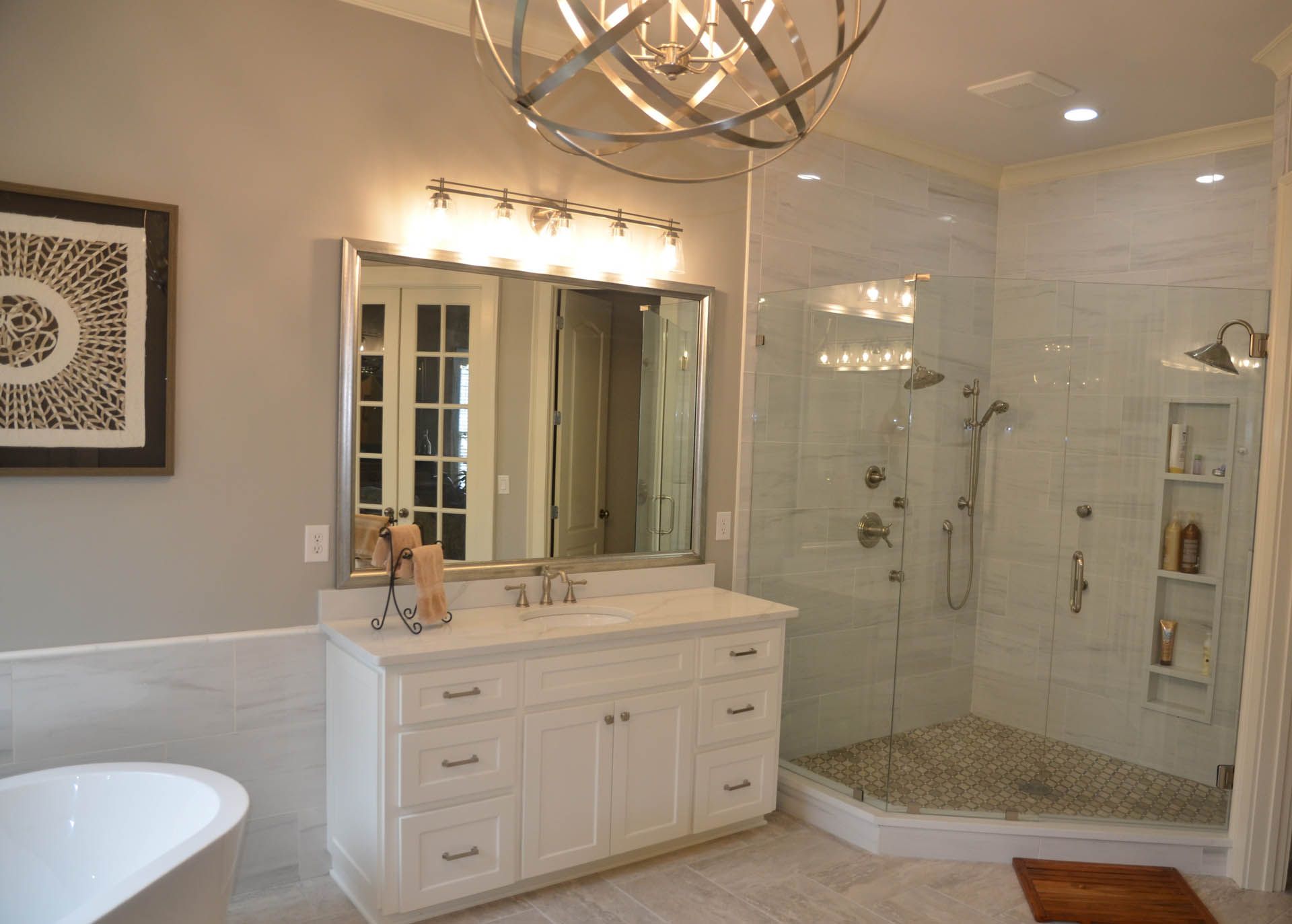 Spacious, modern bathroom with white vanity, glass shower, marble walls, and an orb chandelier.