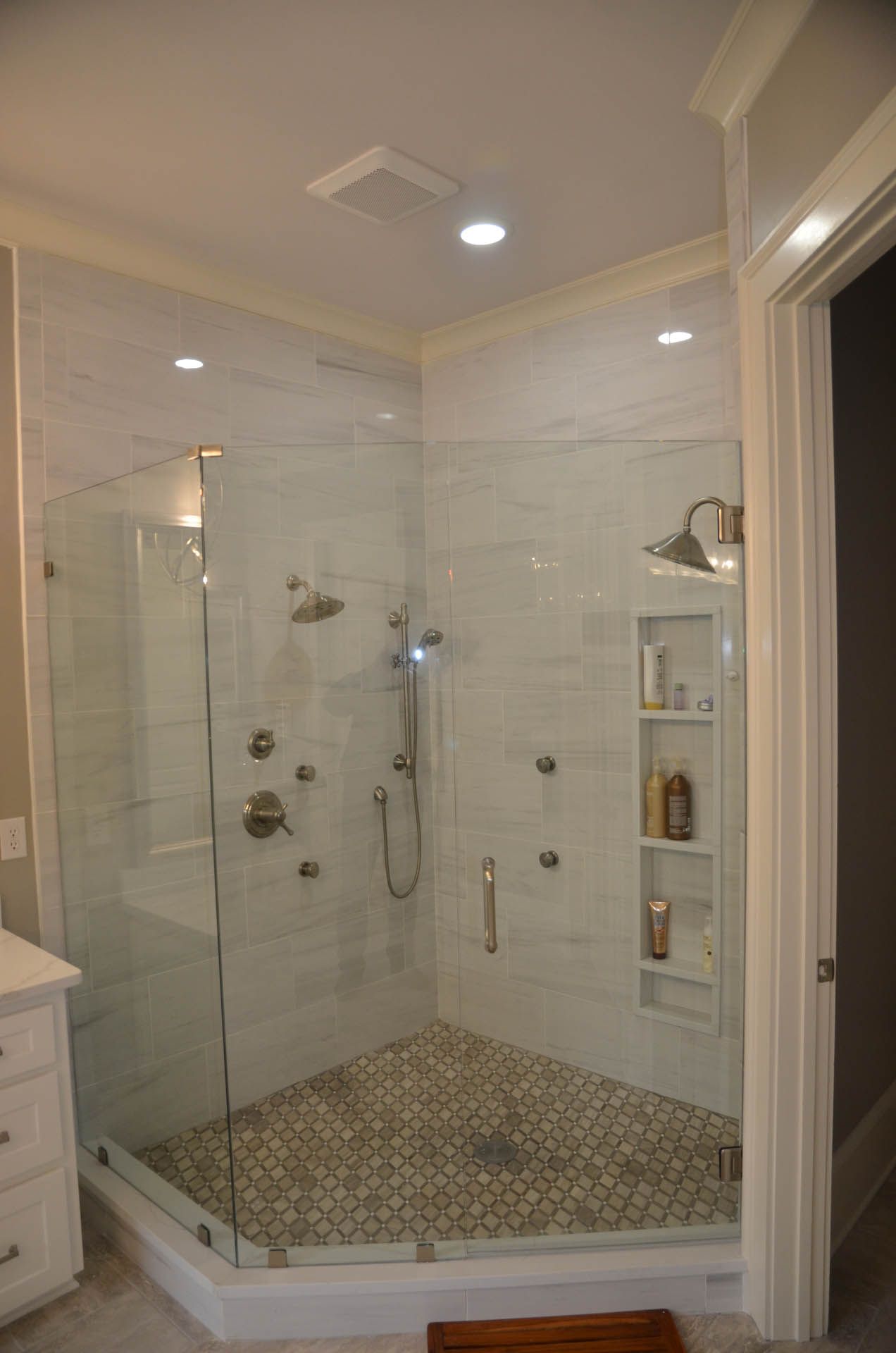A modern, glass-enclosed shower with marble-like tile walls, a tiled floor, and multiple showerheads. A built-in shelf holds toiletries.