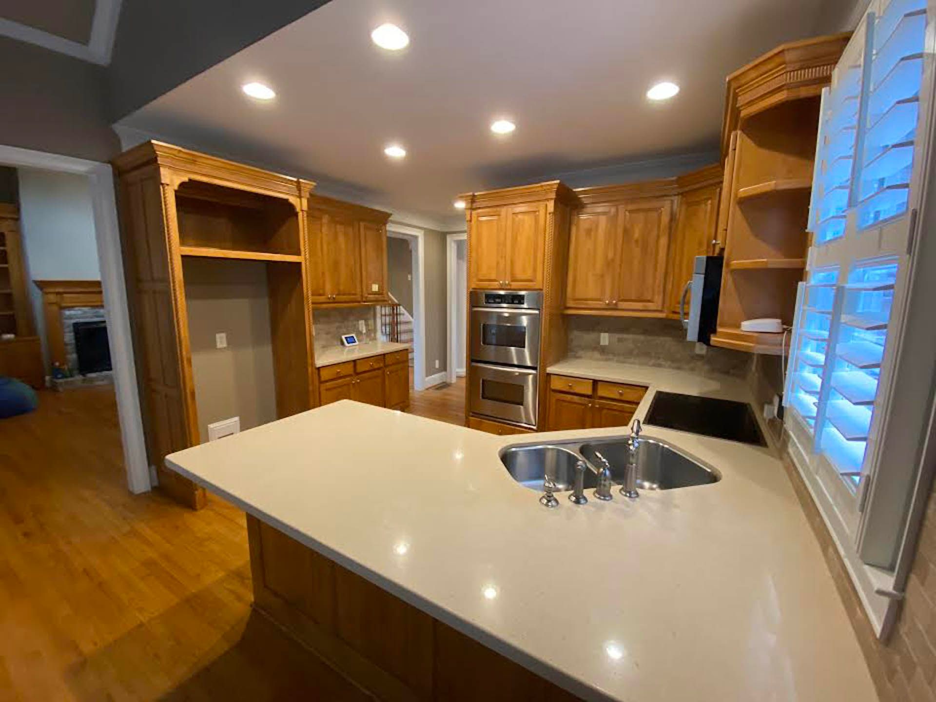 A kitchen with light-colored wooden cabinets, a large island with a sink, and a built-in oven. Natural light streams in from a window.