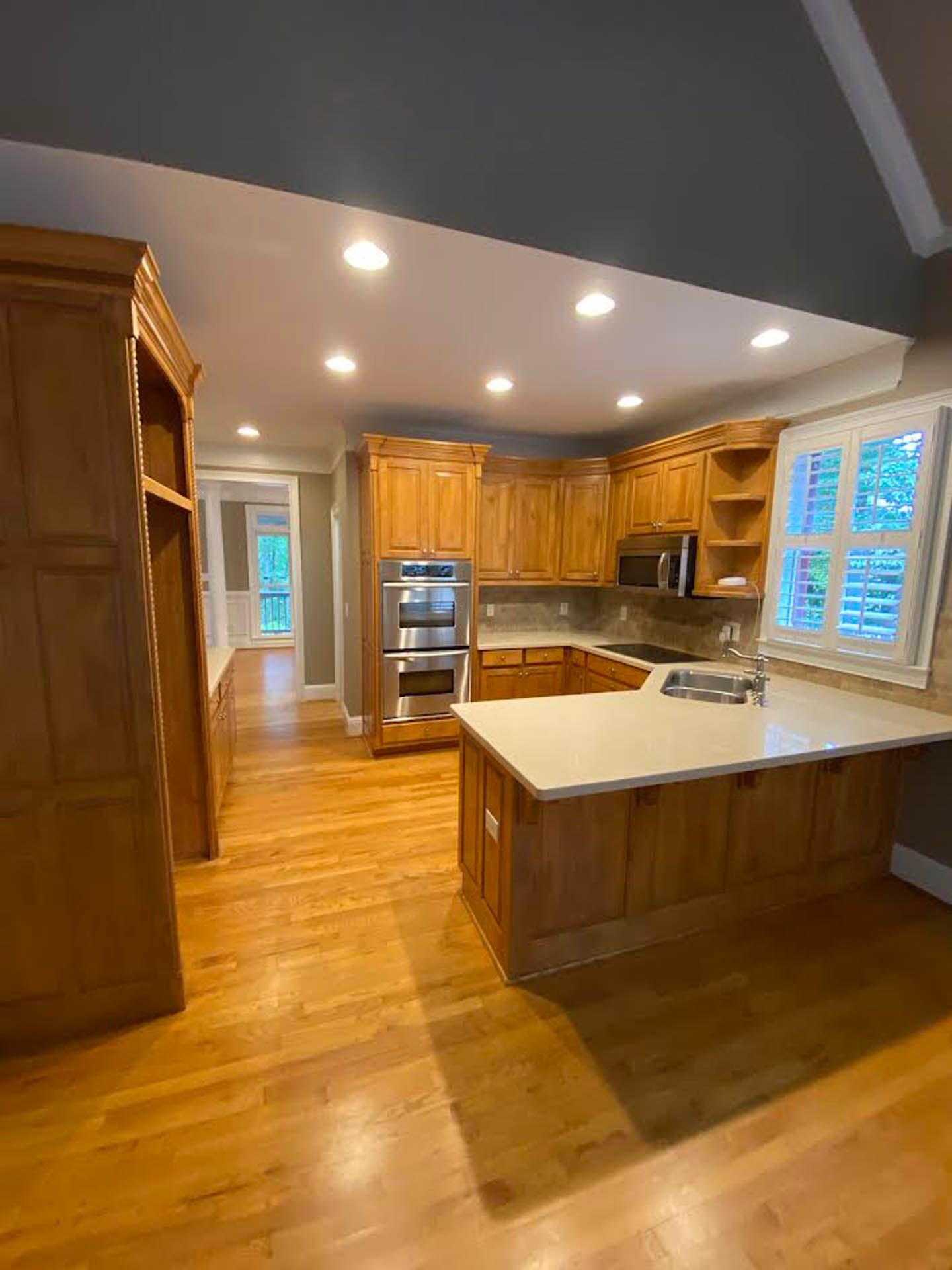 A kitchen with wooden cabinets, light countertops, and hardwood floors. Includes an island, a built-in oven, and a window with shutters.