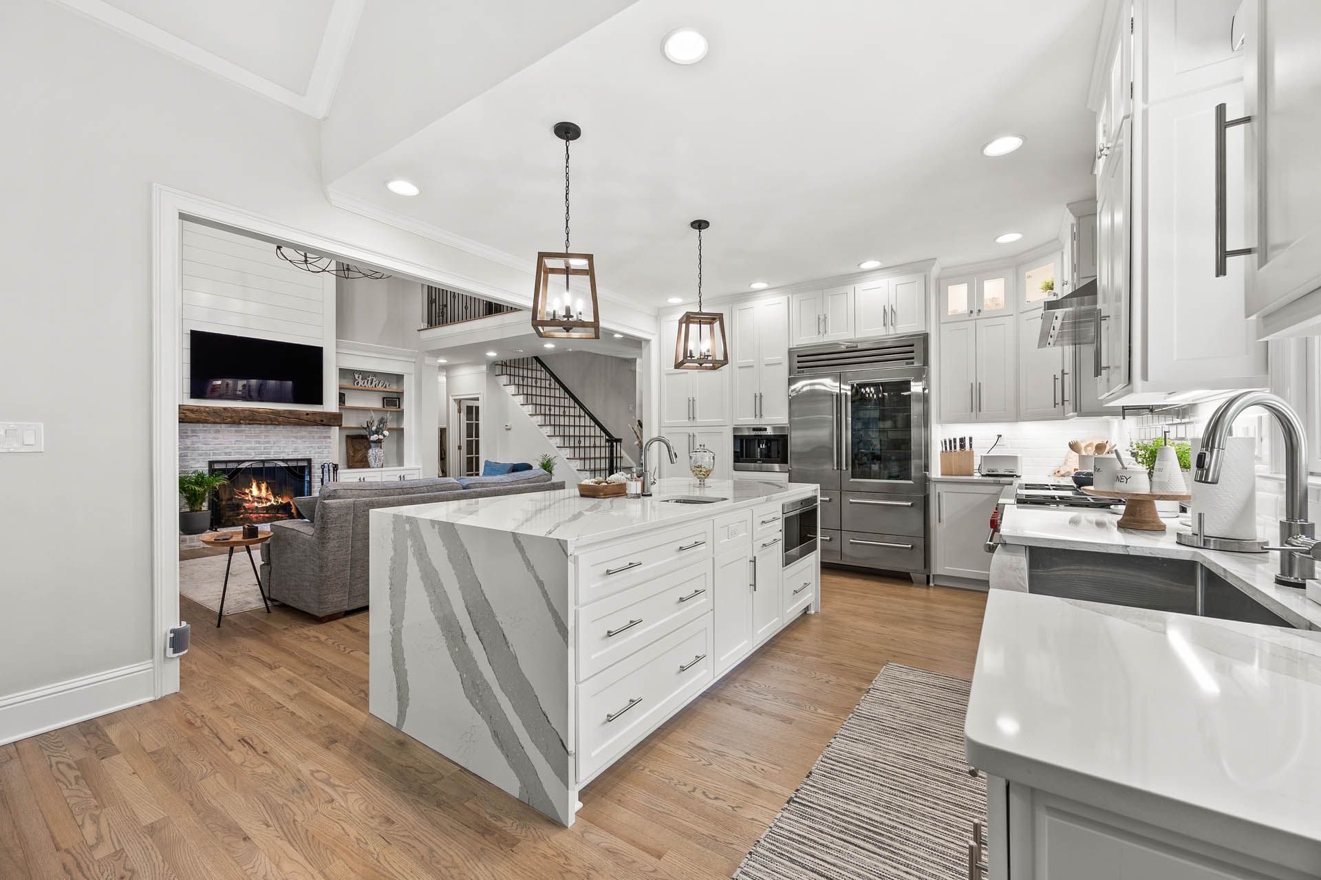 Spacious modern kitchen with white cabinets, a large island, and wood floors. A living area with a fireplace is visible in the background.