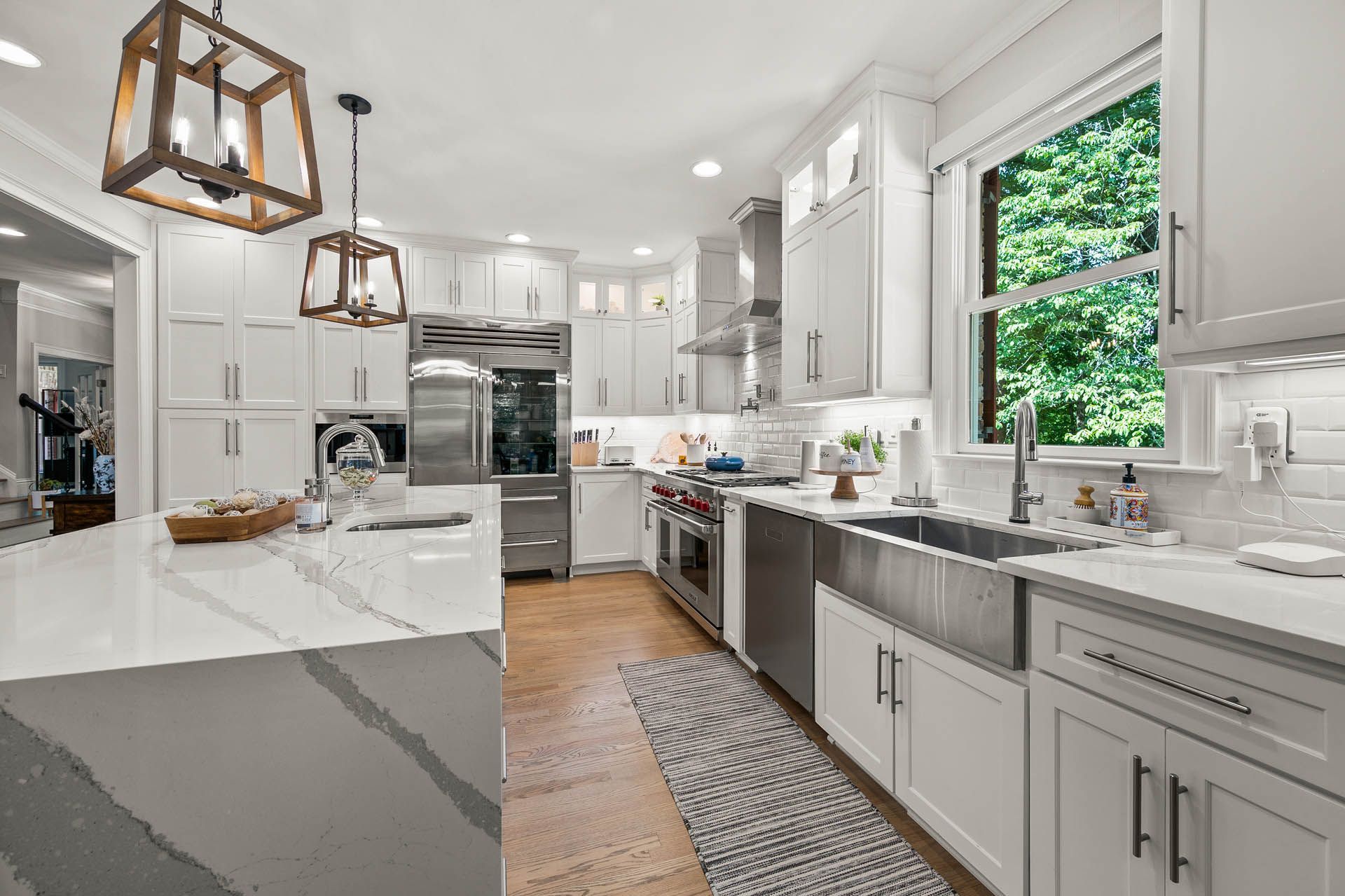 Bright, modern kitchen with white cabinets, stainless steel appliances, and a large island with a gray countertop. A window offers a view of green foliage.