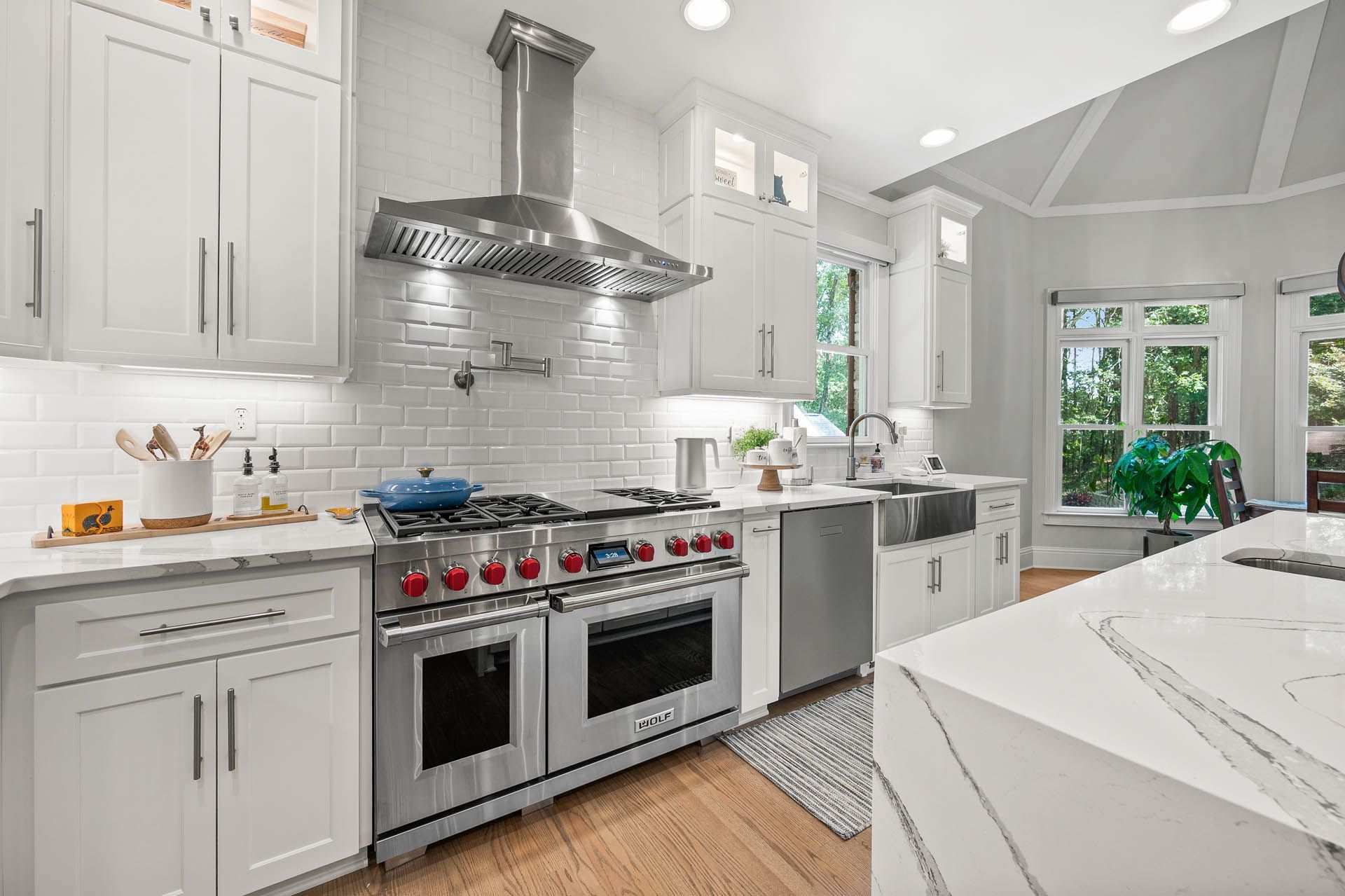 White kitchen with stainless steel appliances, white cabinets, and a large island. A stove, sink, and window are visible.