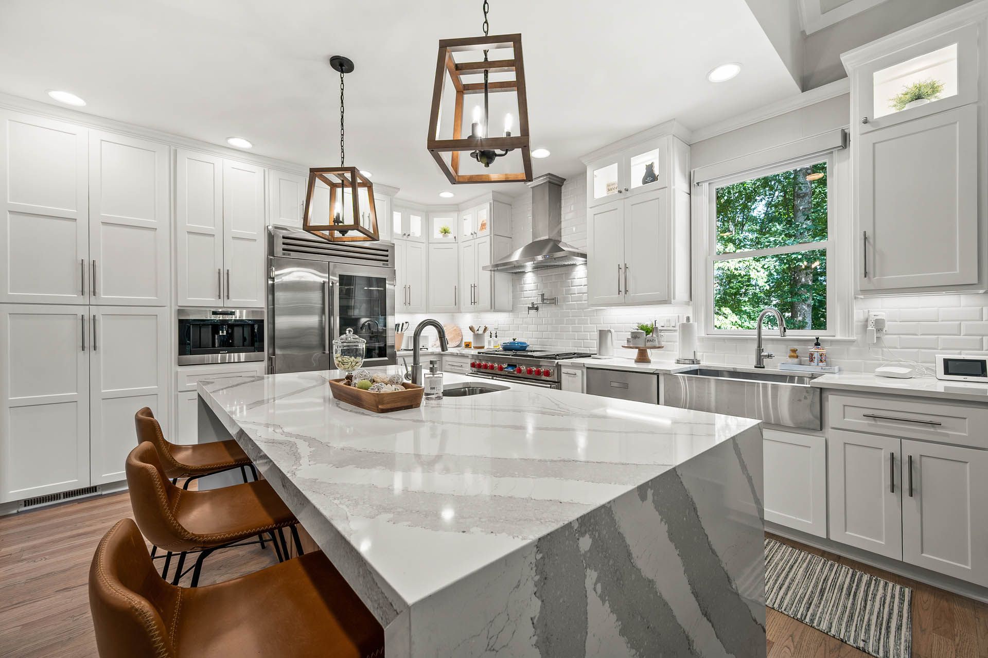 A modern white kitchen with a large island, wooden pendant lights, and stainless steel appliances. Brown leather bar stools sit at the island.