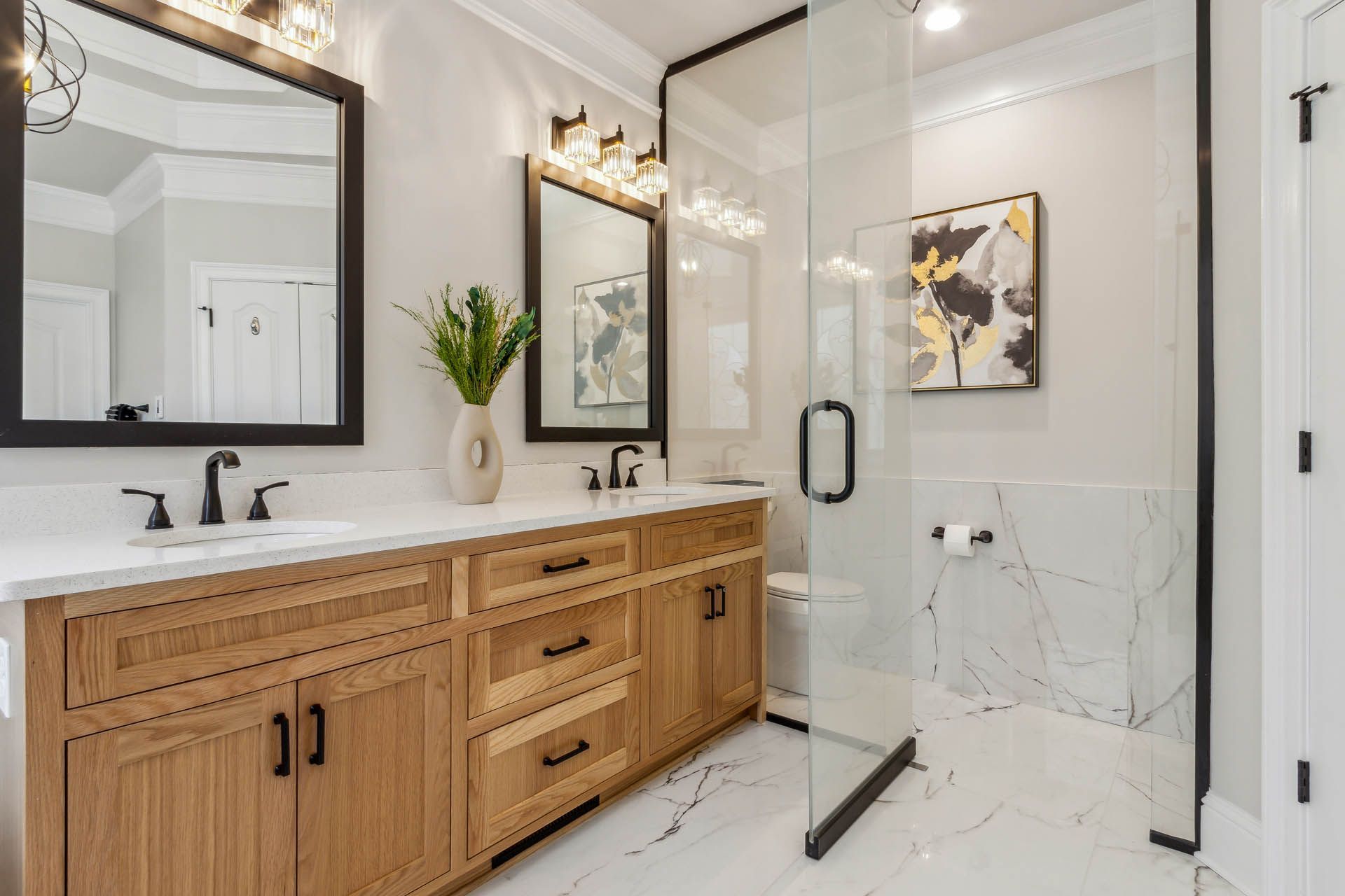 Spacious bathroom with a wooden vanity, marble floors, and a glass-enclosed shower. Two mirrors, light fixtures, and floral art add detail.