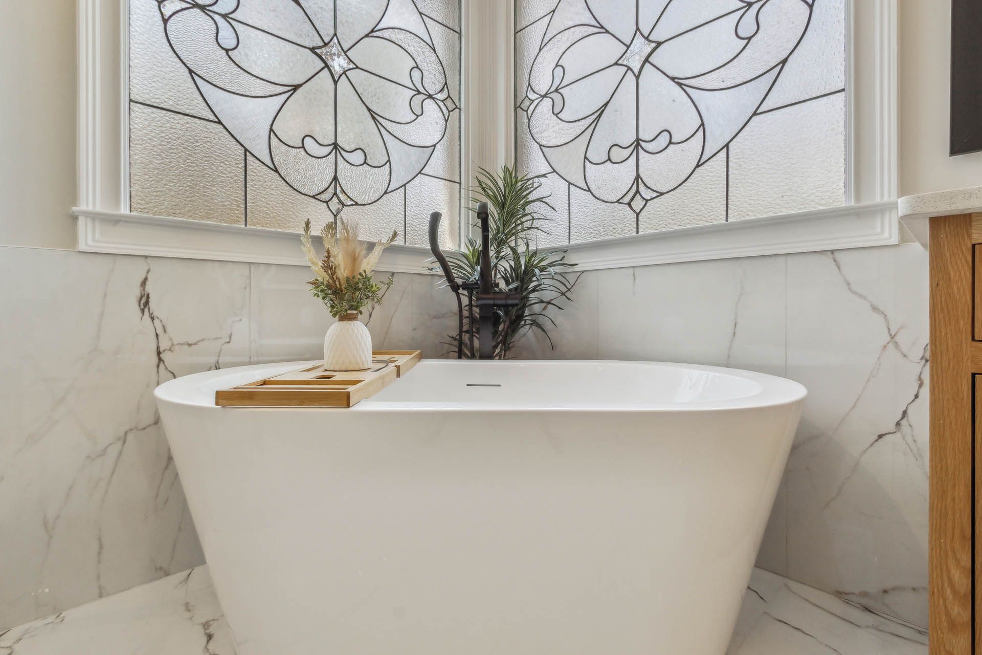 Freestanding white bathtub in a corner with marble walls and a stained-glass window above. A wooden tray holds decor.