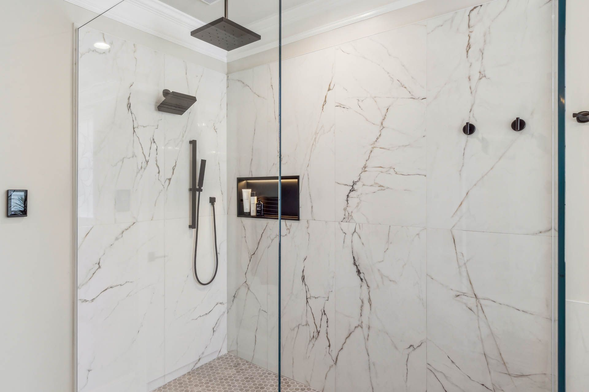 A modern shower with white marble-look tiles, black fixtures, and a glass door. The floor is covered in small, round, light-colored pebbles.