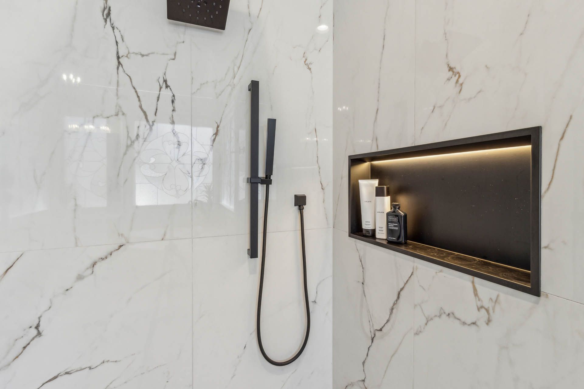 Modern bathroom with white marble tile, a black shower head and wand, and a recessed shelf with toiletries.