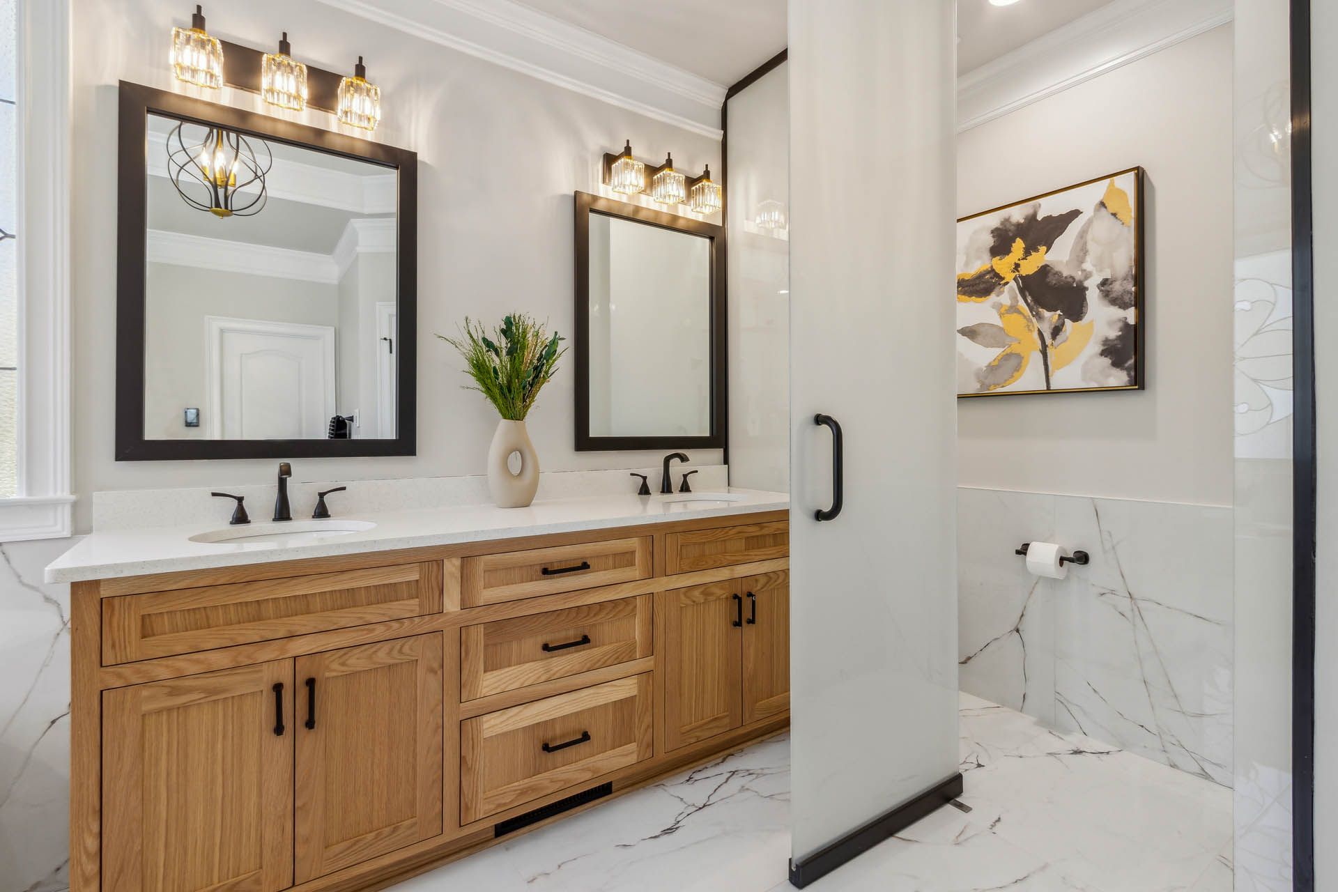 A light-filled modern bathroom with a wood vanity, black framed mirrors, and a white marble-look floor and walls.