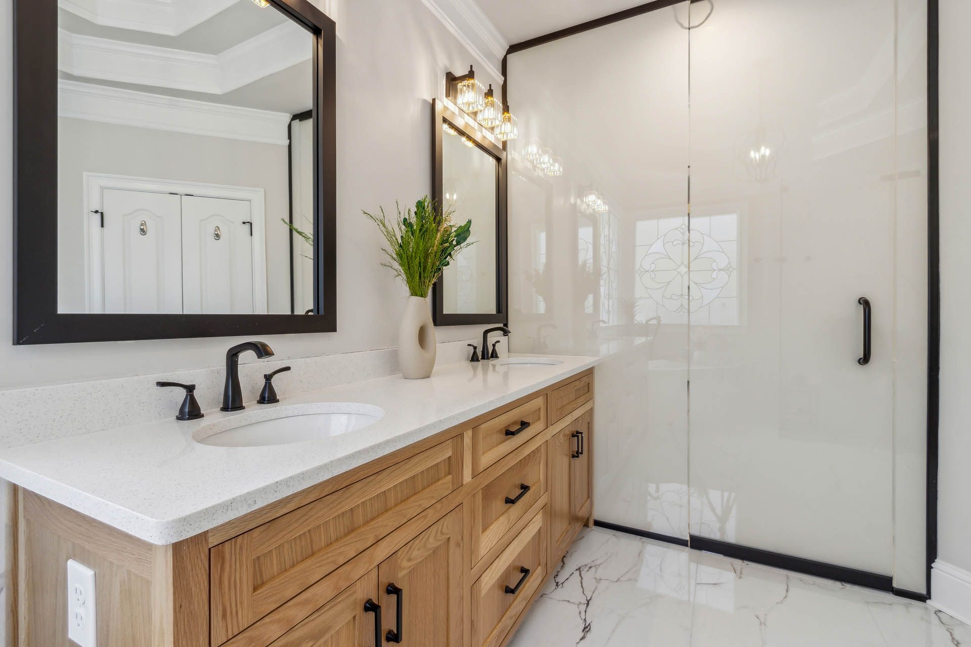 Bathroom with wooden vanity, white countertop, black fixtures, and a glass shower. White walls and a marble-look floor.