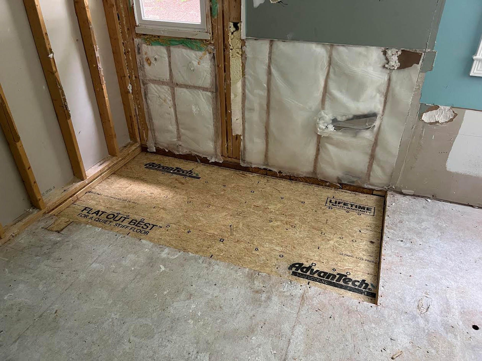 A construction site shows a bare plywood floor with a wooden frame, surrounded by damaged walls and insulation.