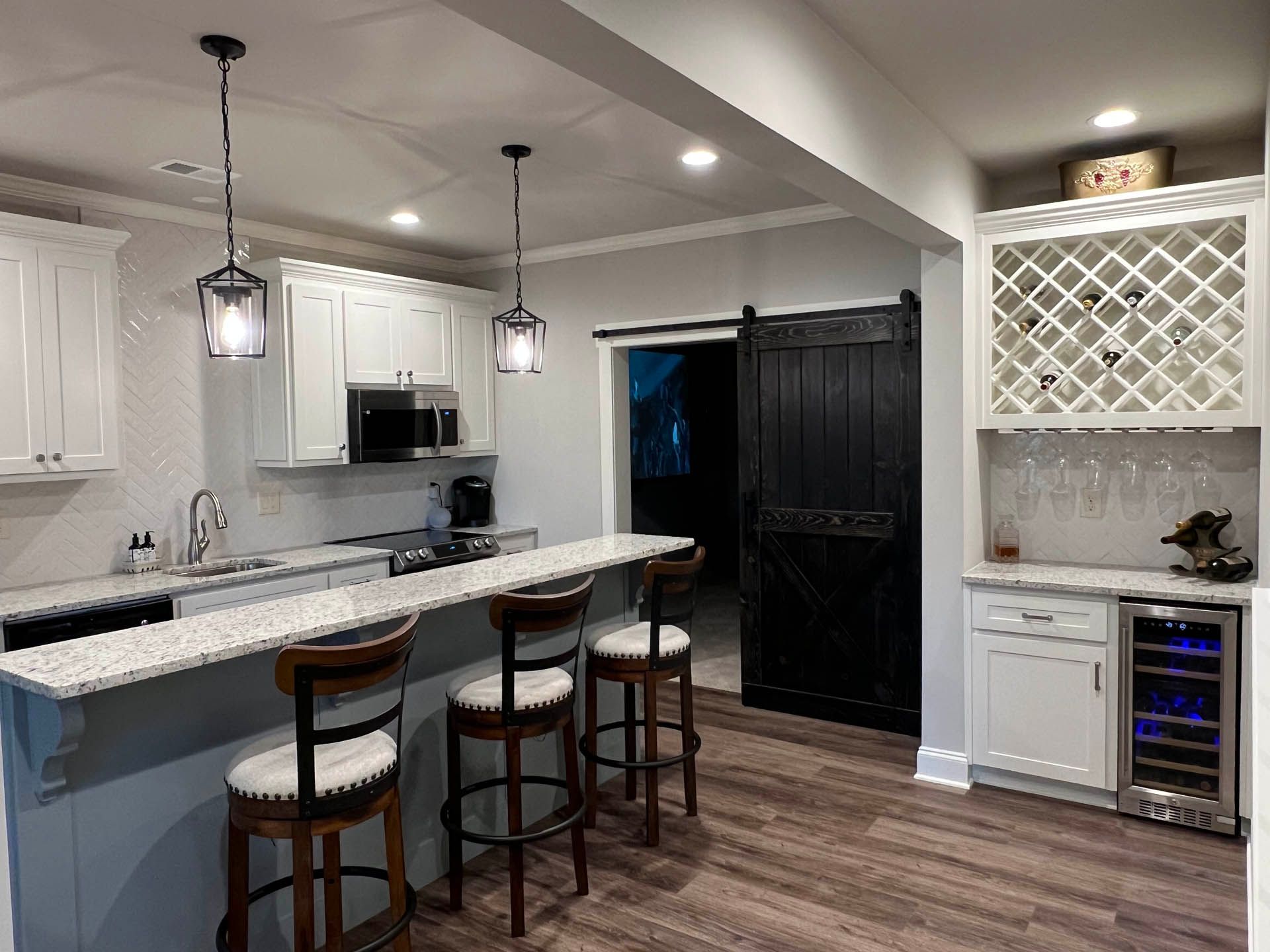 A modern kitchen with a breakfast bar, white cabinets, and a black barn door. Features include a wine rack and a wine fridge.