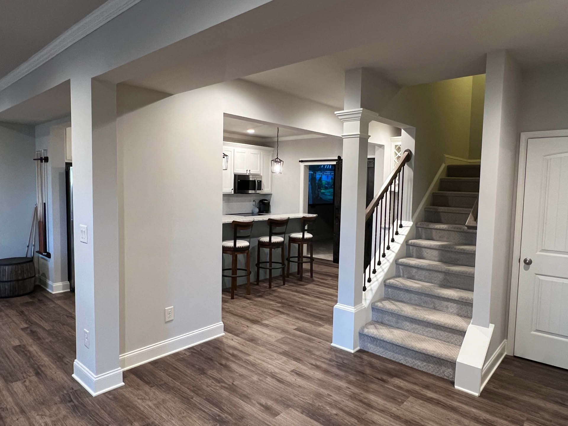 Interior view of a finished basement with a kitchen, staircase, and columns.  Gray walls, wood-look flooring, and white trim throughout.