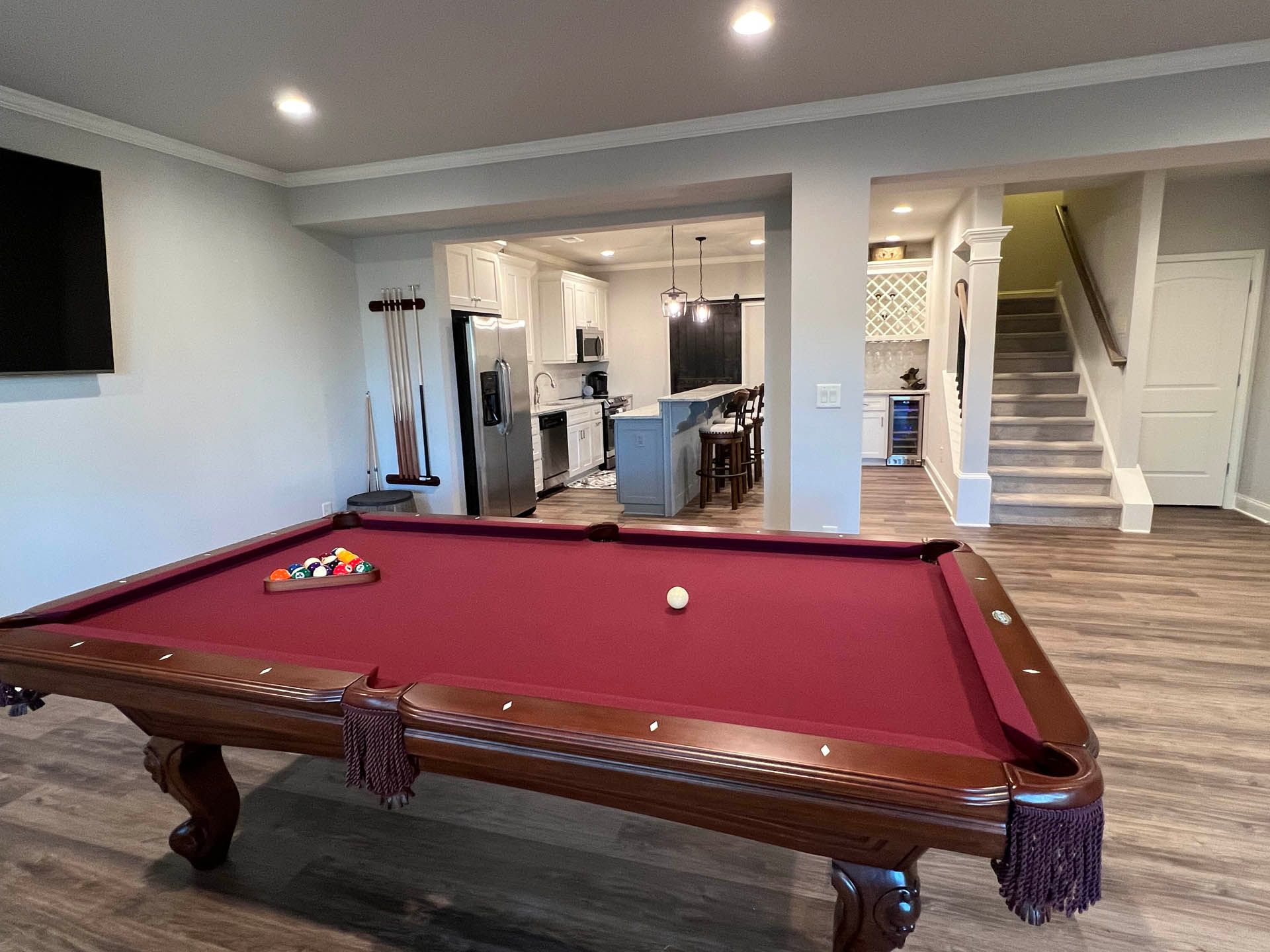 A pool table with red felt is in a game room. Through an opening, a kitchen and stairs are visible.