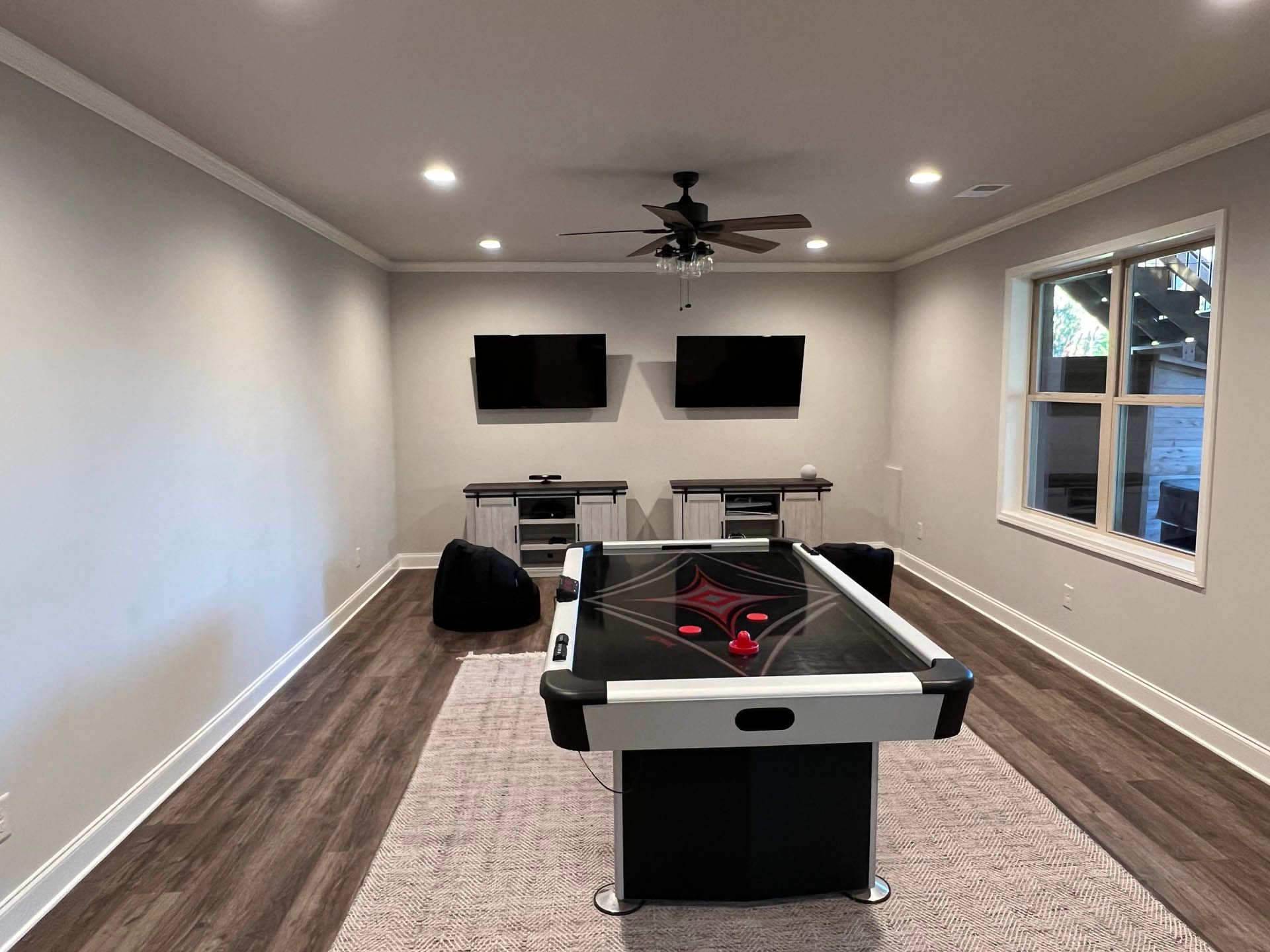 A game room with an air hockey table, two TVs on the wall, and a window. The room has gray walls and brown flooring.